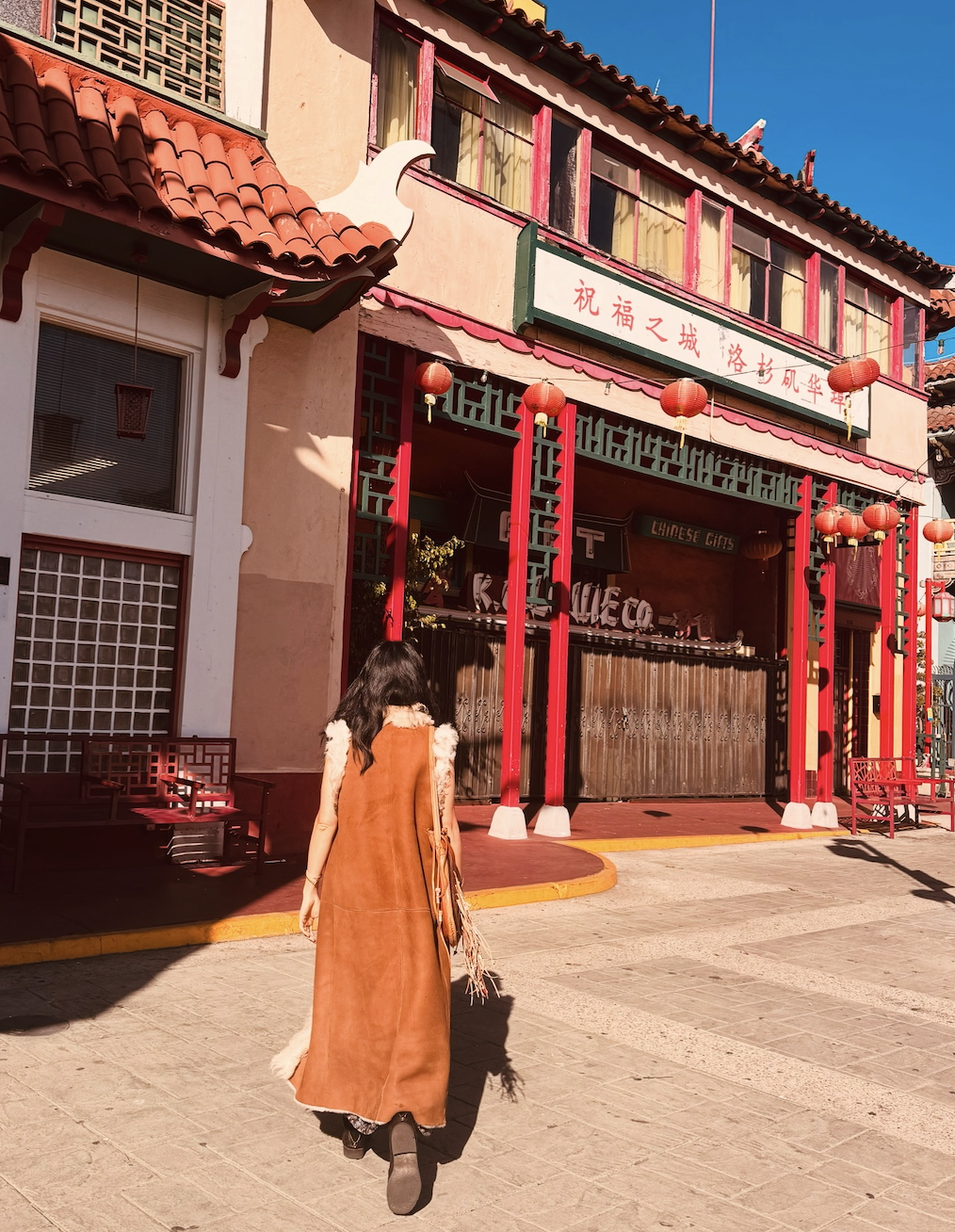 A person walking towards a Chinese-themed building decorated with red lanterns and traditional architecture, with a bright blue sky overhead.
