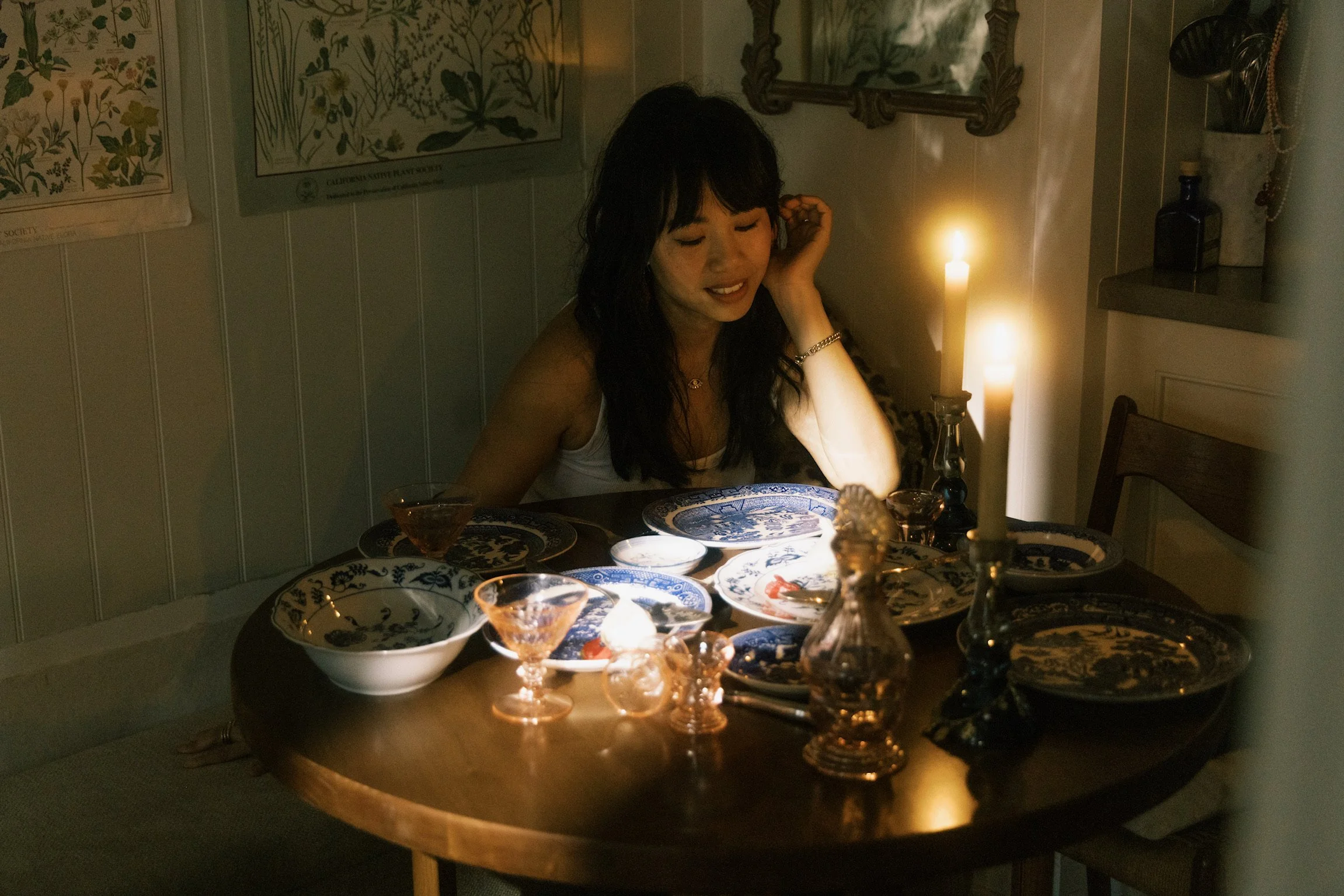 A woman with long dark hair sitting at a dining table illuminated by candlelight, with blue and white patterned dishes and glassware, in a cozy room decorated with botanical prints and wooden furniture.