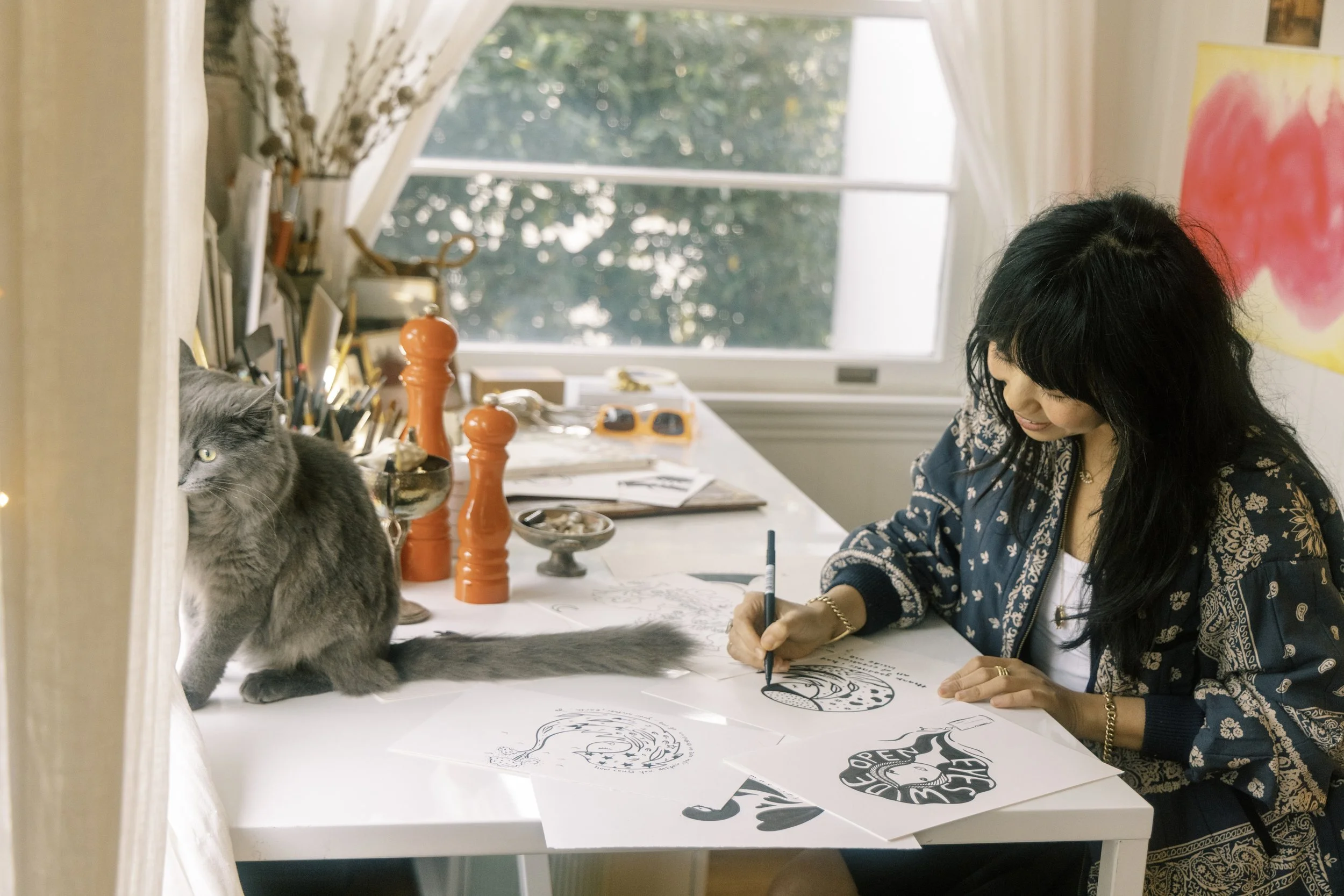 A woman with black hair drawing sketches at a desk in a room with a window, next to a gray cat, with art supplies and drawings on the desk.