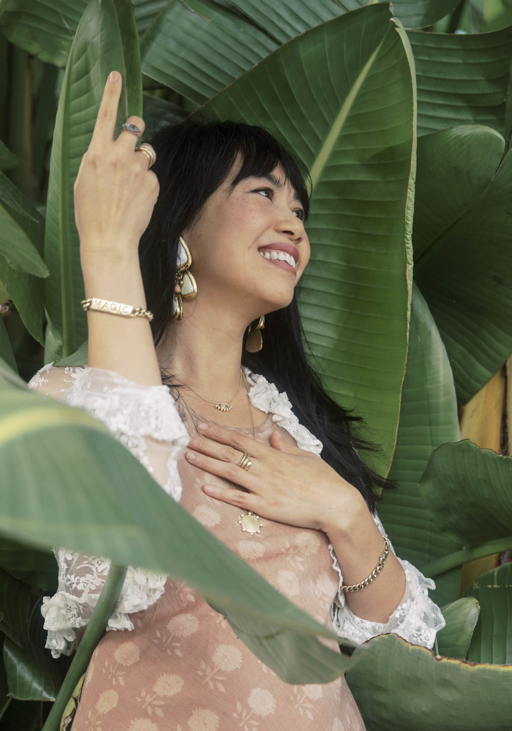 A smiling woman with dark hair, wearing jewelry, standing among large green leaves, touching her chest with her right hand, and showing a peace sign with her left hand.