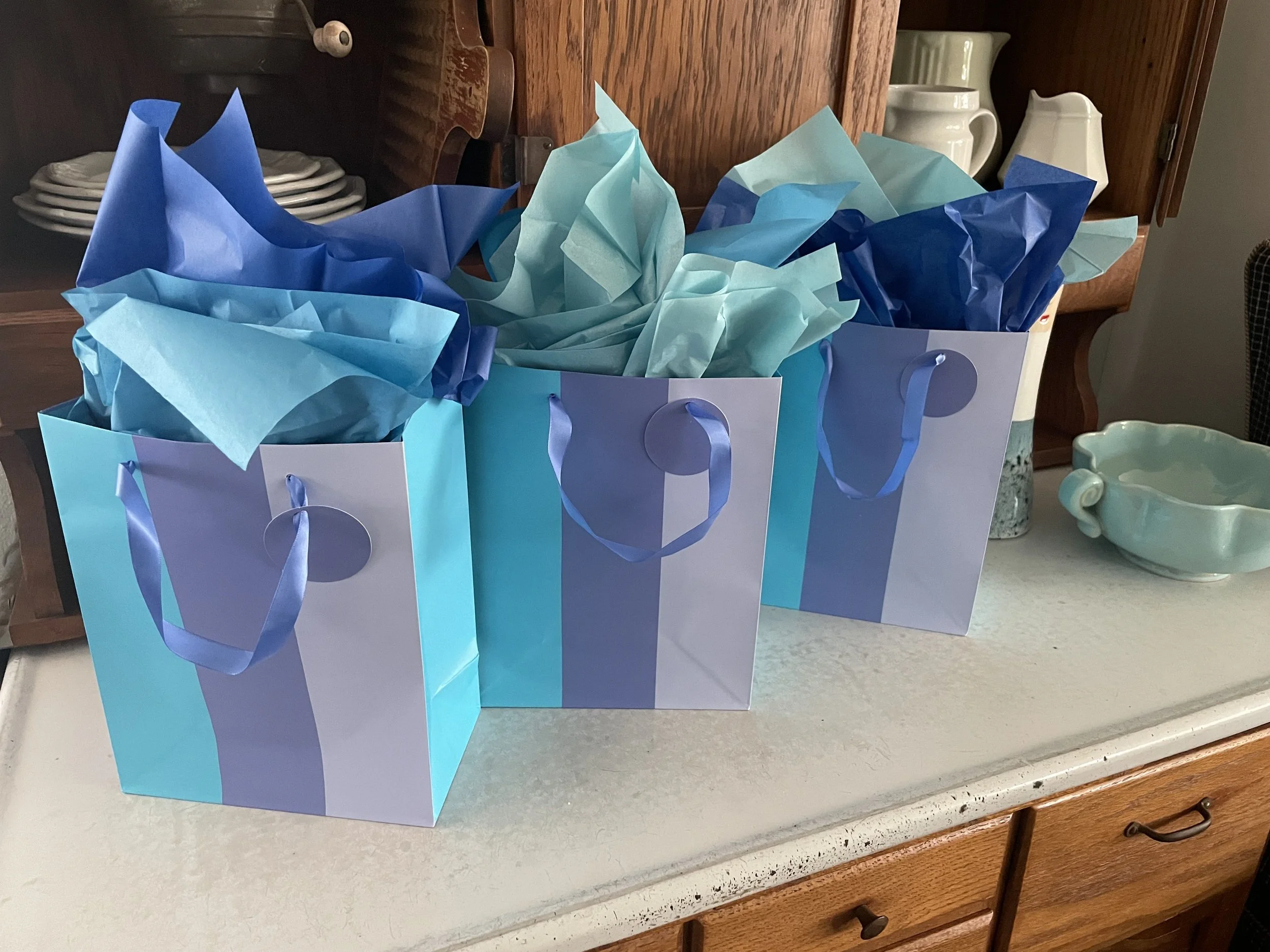 Beginners American Mahjong event at Scripps Ranch, San Diego. Three gift bags with blue stripes, filled with blue tissue paper, placed on a countertop with decorative items in the background.