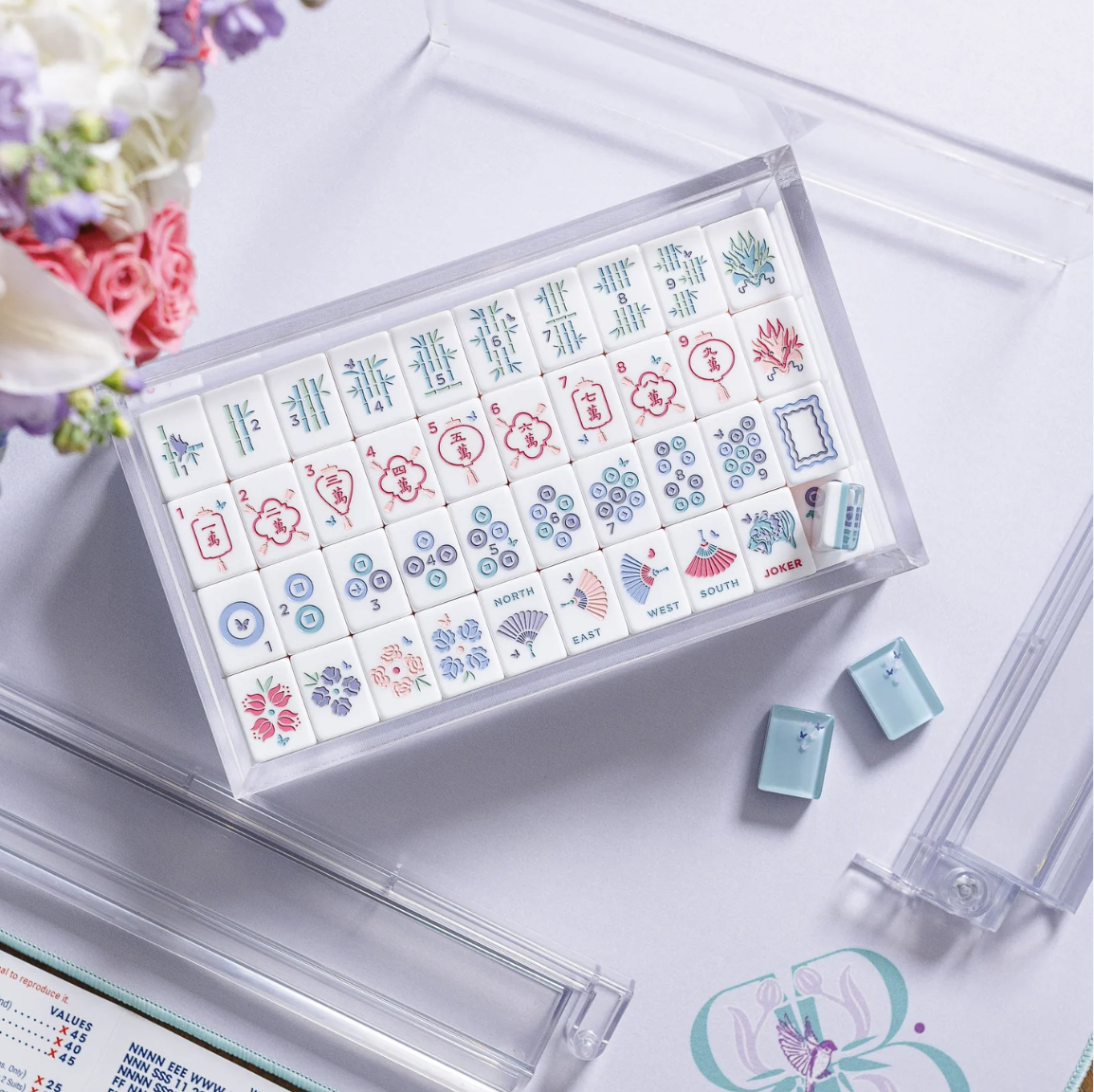 An open box of Mahjong tiles with various designs, including bamboo, circles, and character suits. There are also wind and dragon tiles, along with flower tiles. The set includes clear racks and is placed on a light surface with flowers nearby.