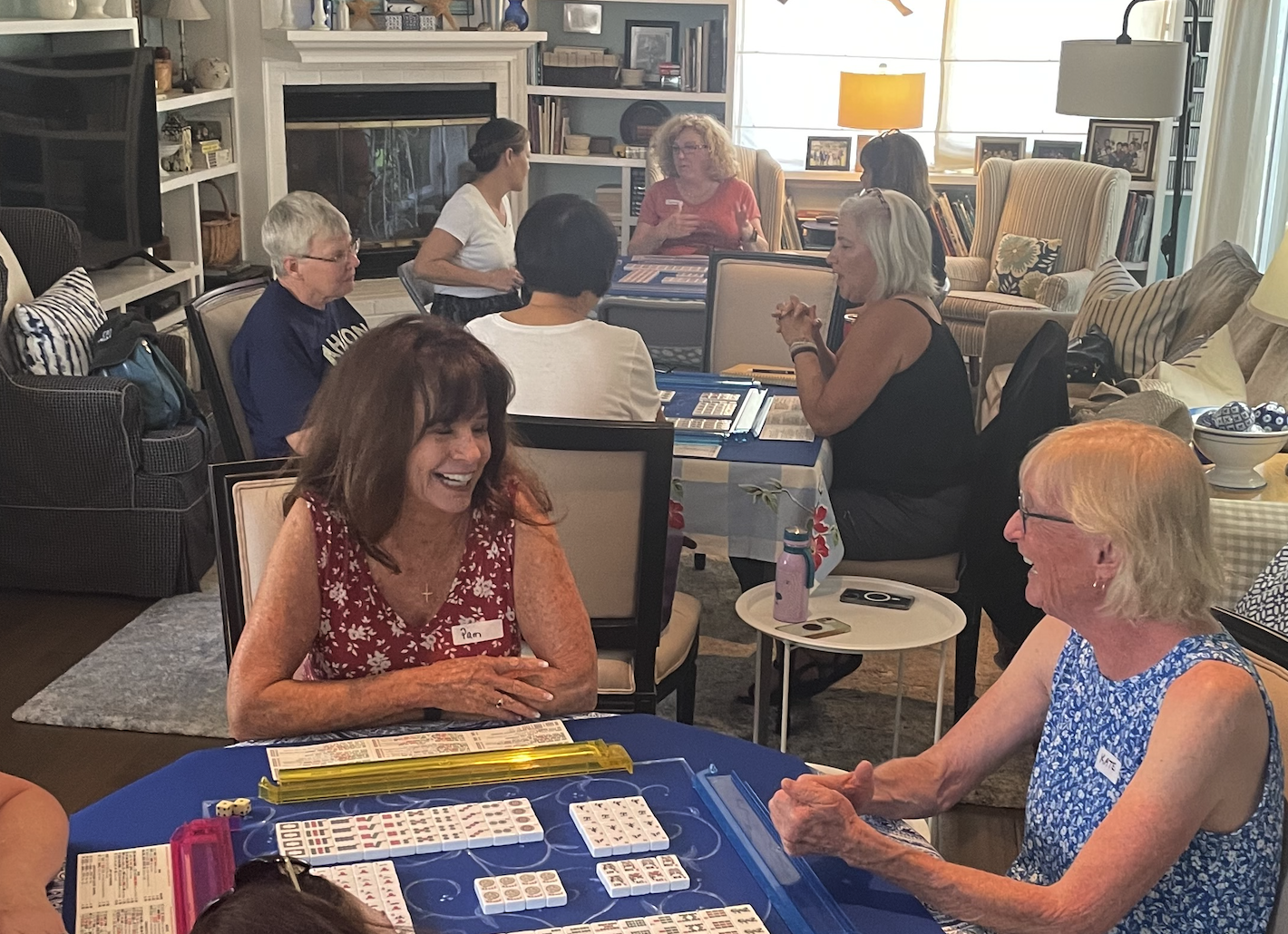 Beginners American Mahjong class at Scripps Ranch, San Diego. Three tables of students playing mahjong in a cozy living room with bookshelves, armchairs, and large windows.