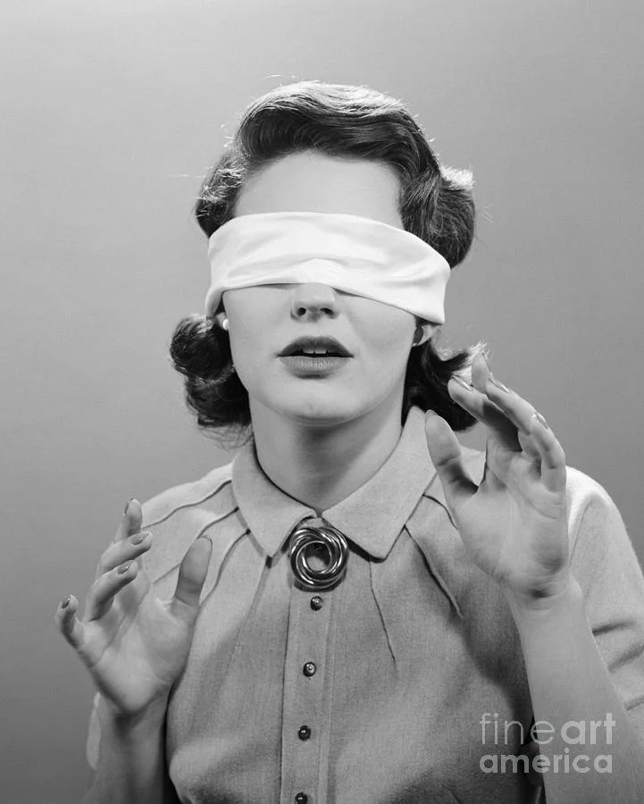 Young woman in 1950s style blouse, also wearing a blindfold, with her hands in front of her.