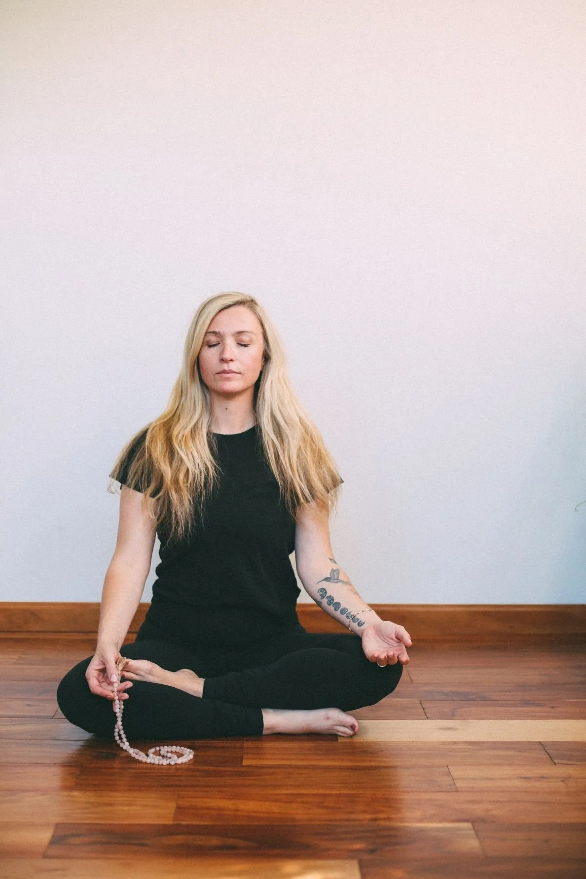 megan cannon in black yoga outfit sitting on wooden floor holding pink mala in right hand