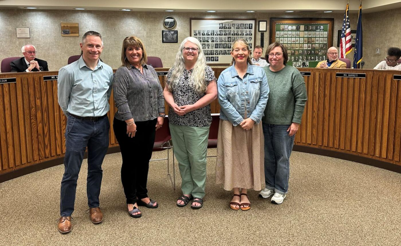 “Five people stand in a row at the front of a meeting room, with a panel of seated individuals behind them and an American flag in the background. The room has wood paneling and a formal setting.”