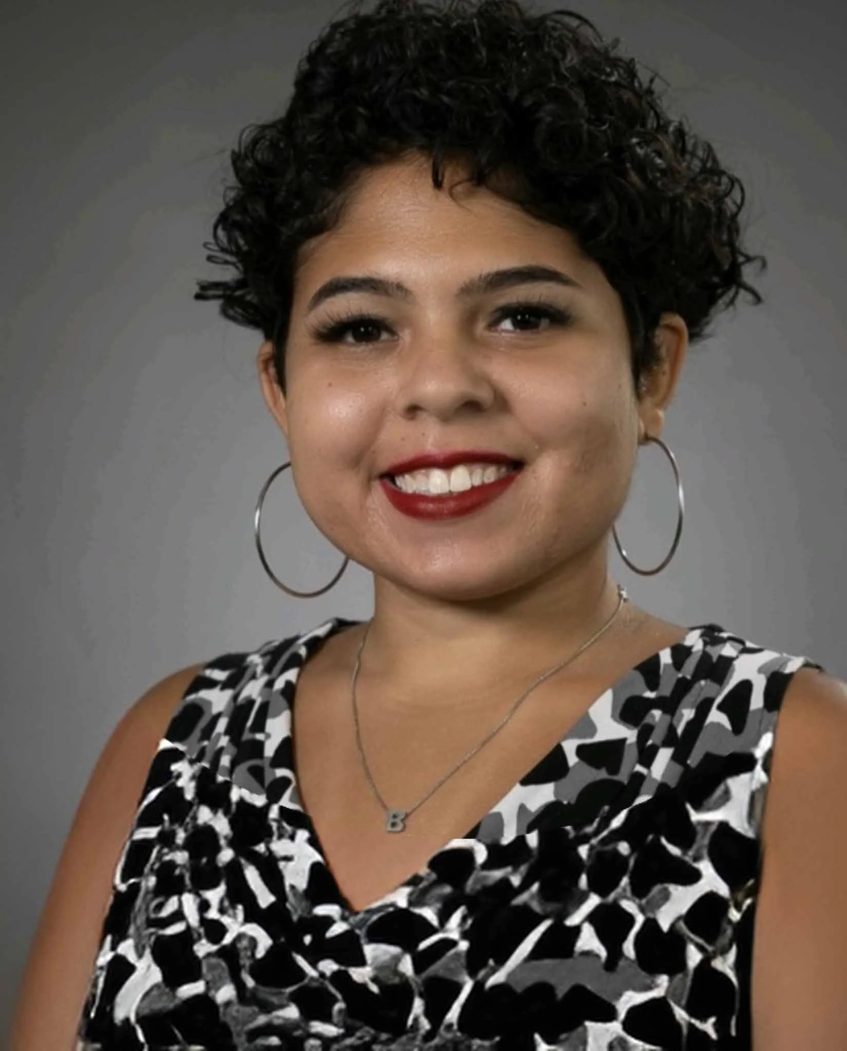 A woman with short, curly black hair smiling, wearing large hoop earrings, a silver necklace with a 'B' pendant, and a black and white animal print sleeveless top against a gray background.