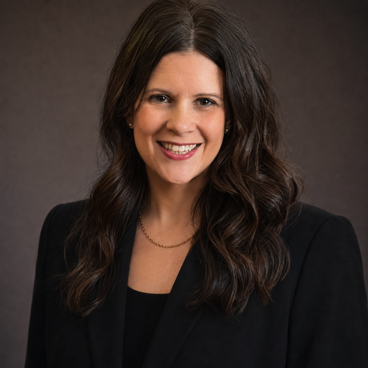 Headshot of a woman with shoulder-length brown hair, smiling, wearing a black blazer and pearl earrings against a gray background.
