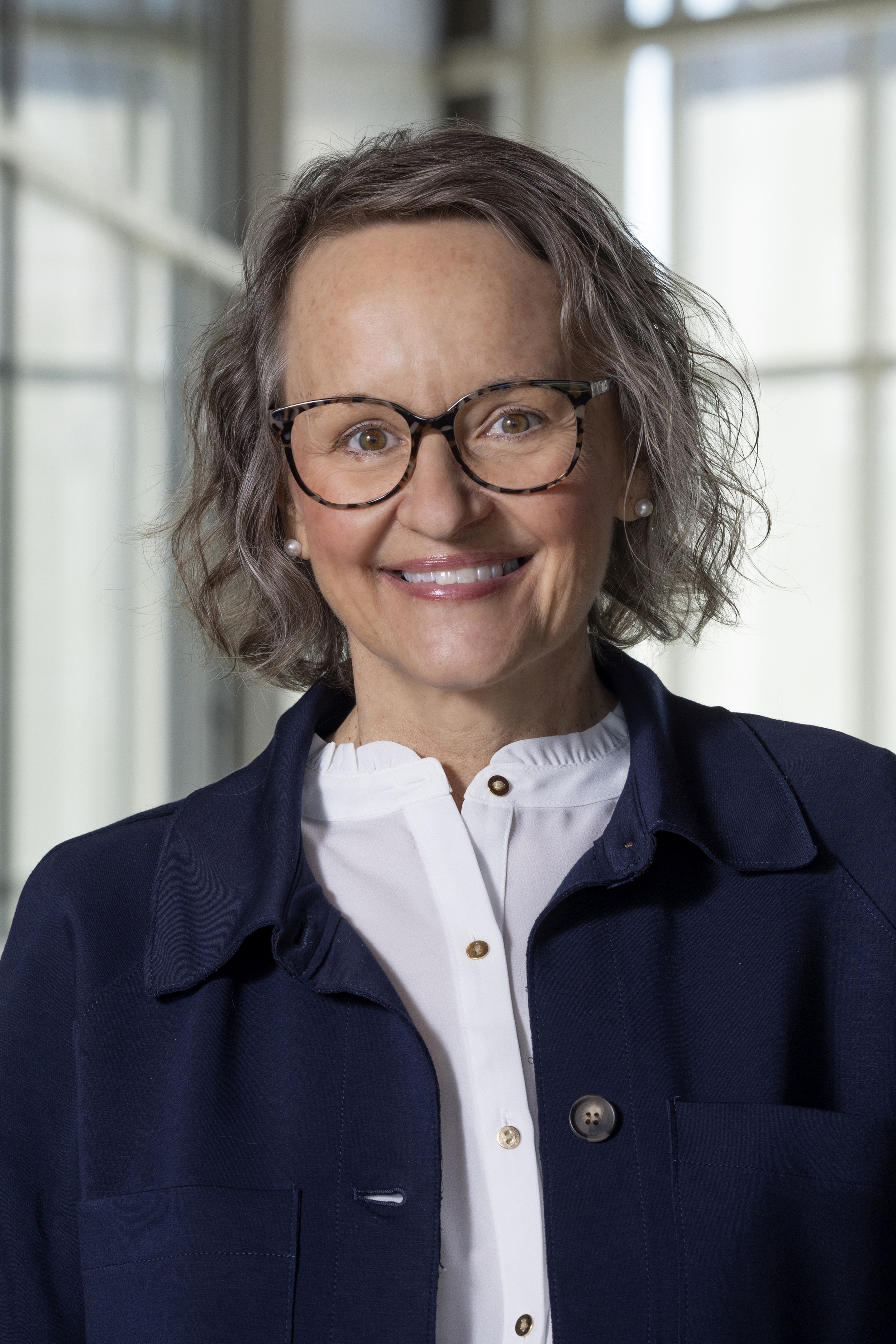 A woman with short brown hair, glasses, and pearl earrings smiling outdoors with a blurred green background.