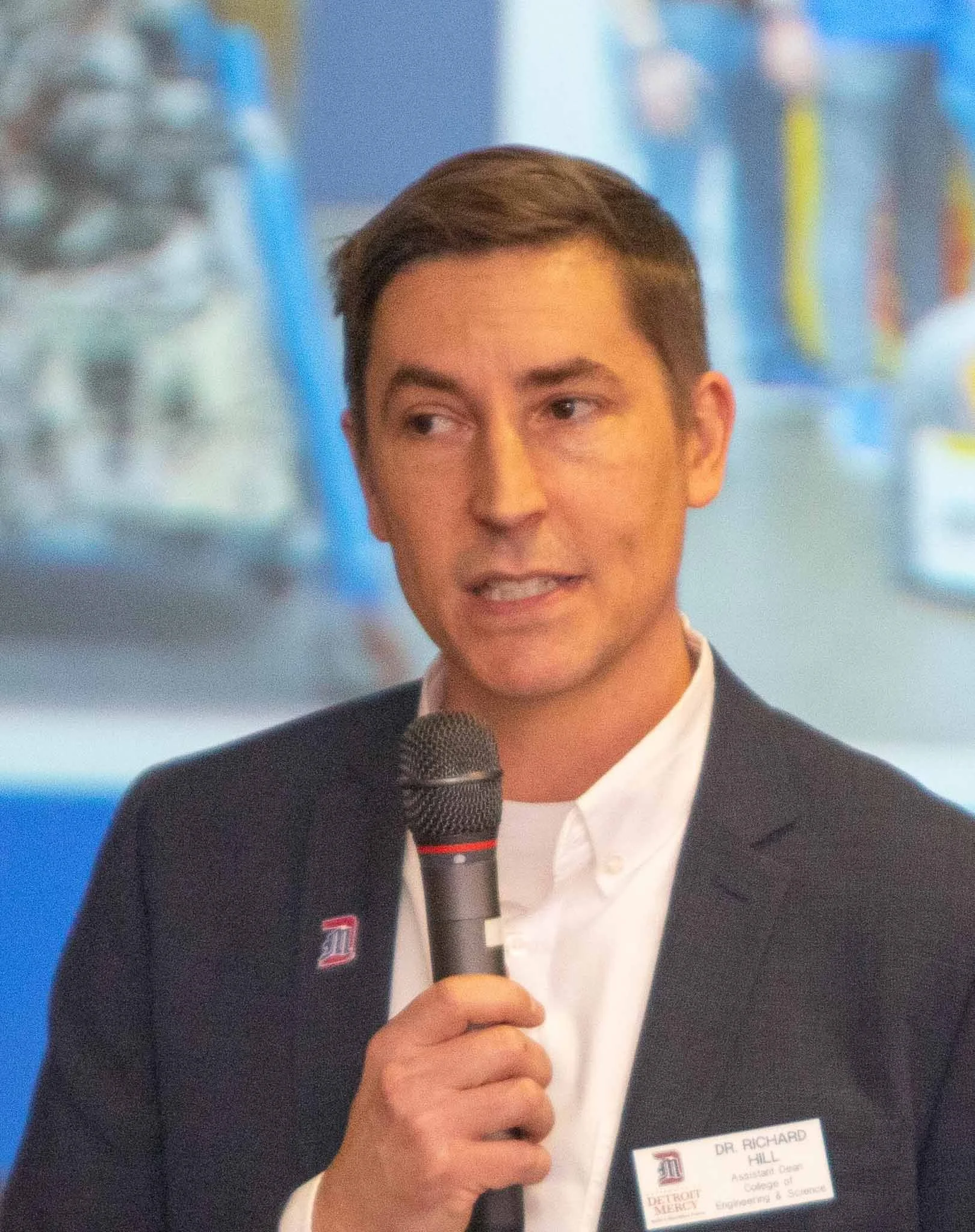 A man in a professional suit holding a microphone, speaking at an event, with a name tag that reads Dr. Richard Hill.