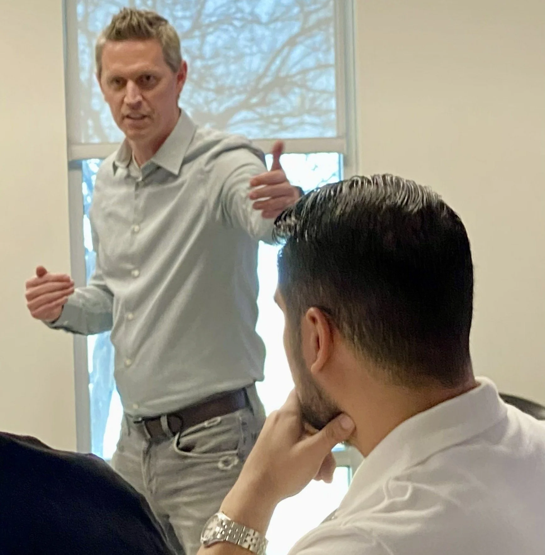 “A person standing at the front of a room gestures with one arm extended while another person sits nearby, looking toward the speaker. A large window in the background shows tree branches outside.”