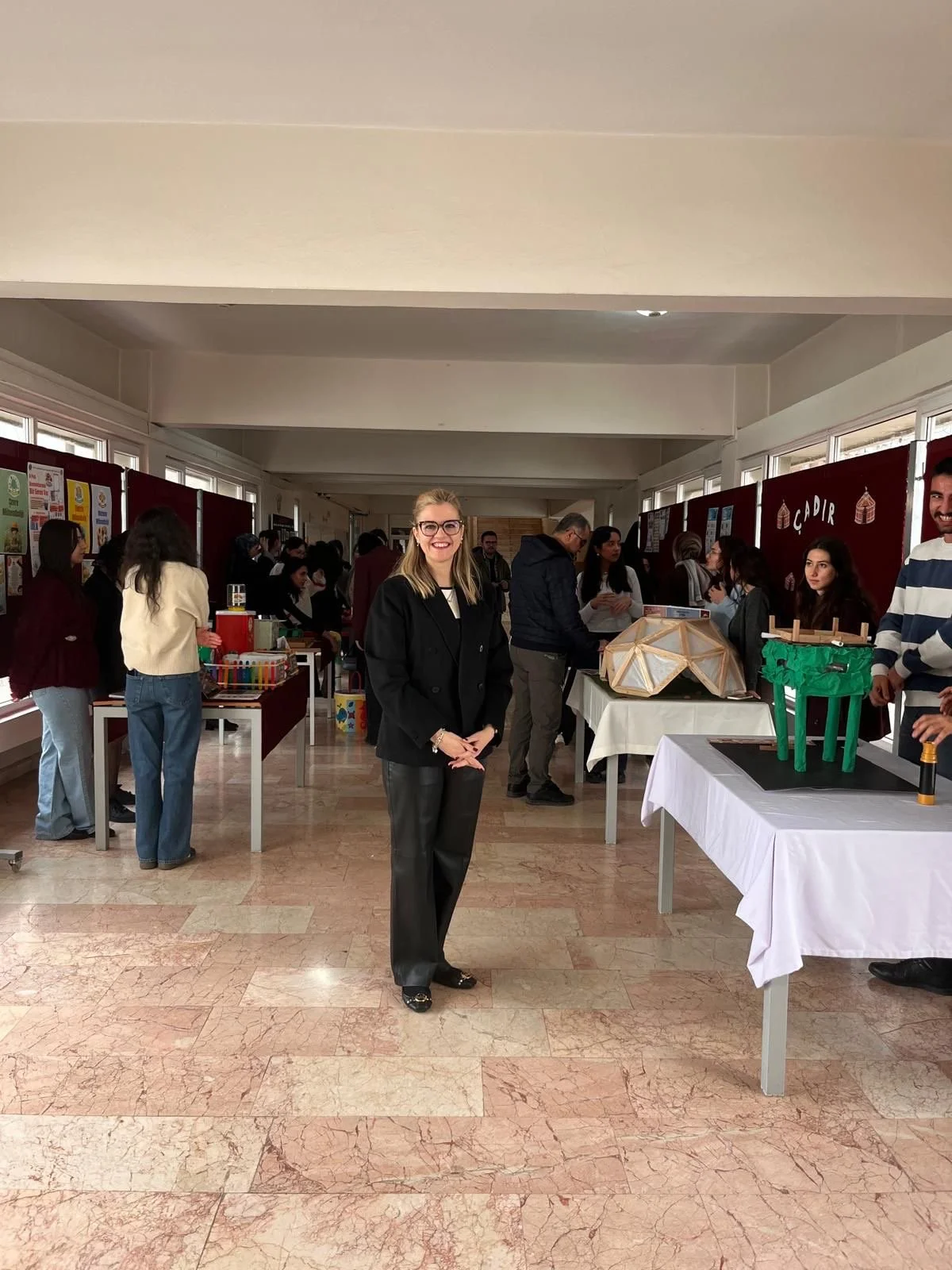 “A person stands in the middle of a large indoor exhibition space where multiple tables display projects and models. Groups of people are gathered around the room viewing the exhibits.”