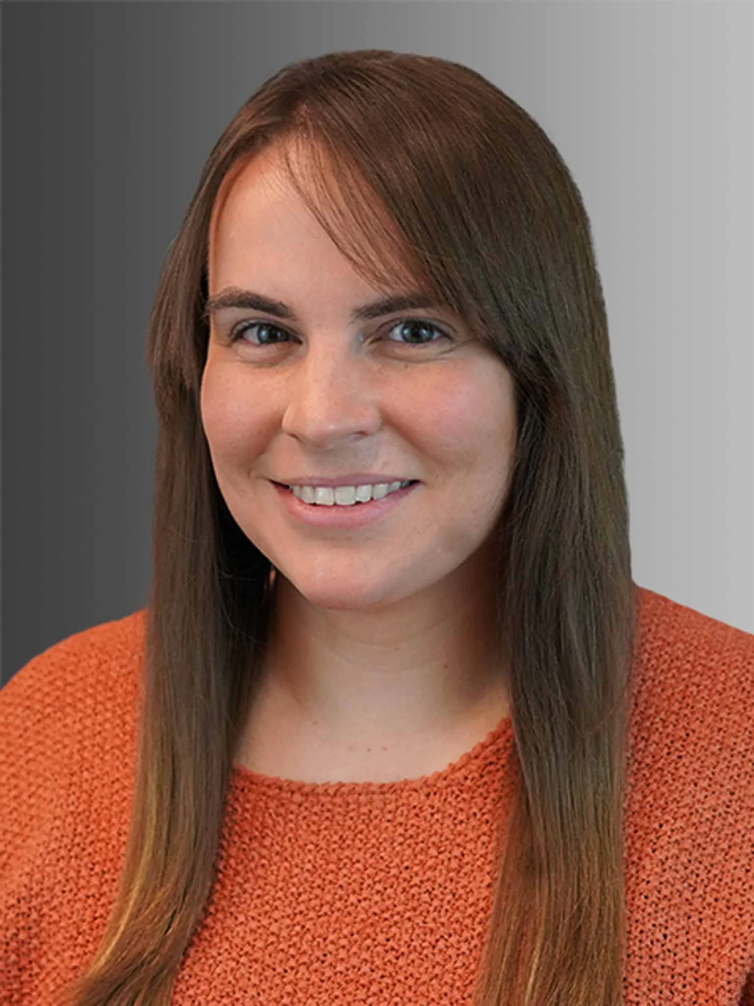 A woman with straight brown hair, smiling, wearing an orange textured sweater, against a gray background.