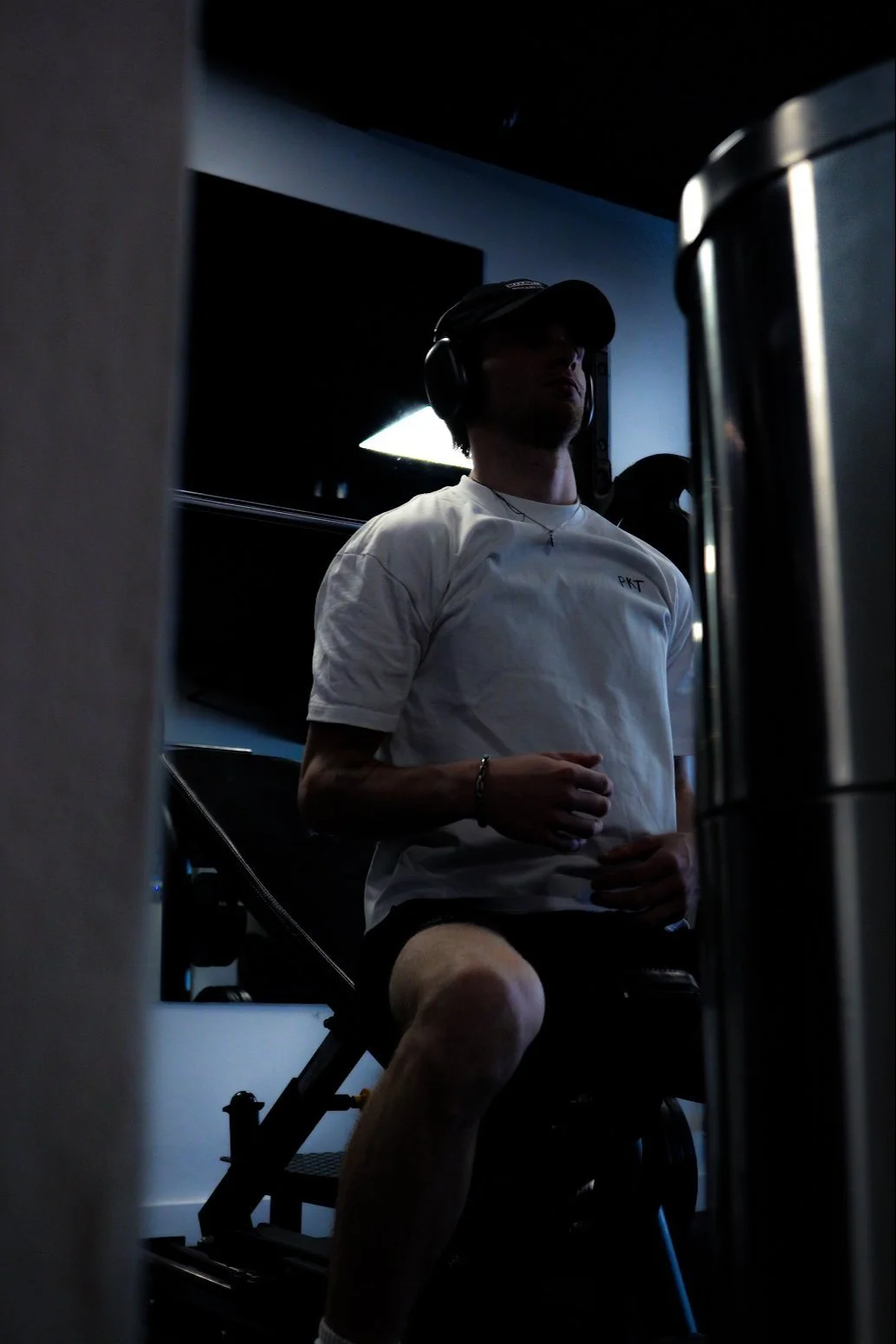 Person in gym wearing headphones and a white t-shirt, seated on exercise equipment.