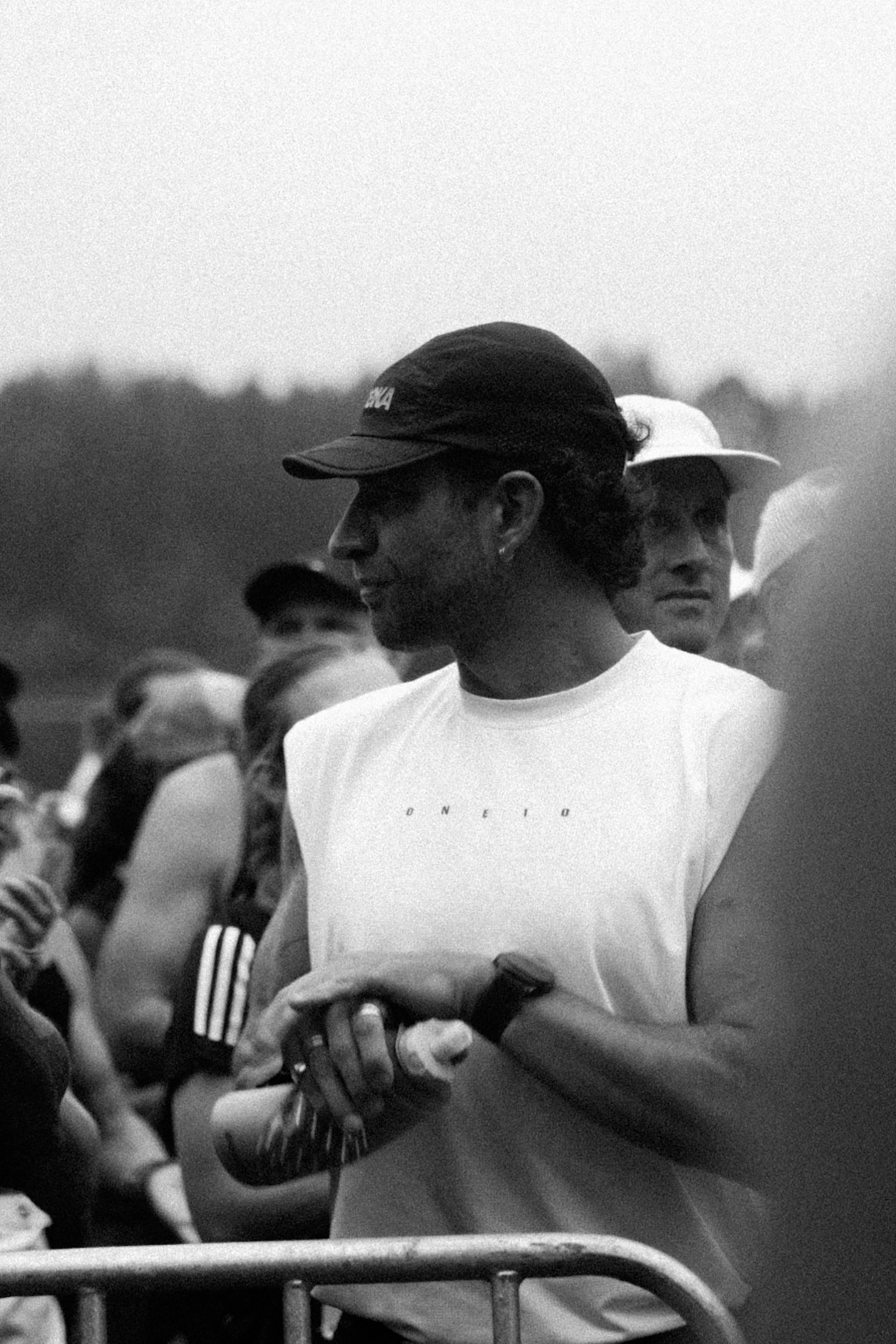 Black and white photo of man in a cap and t-shirt with the word 'Oneten' on it, surrounded by people in hats.