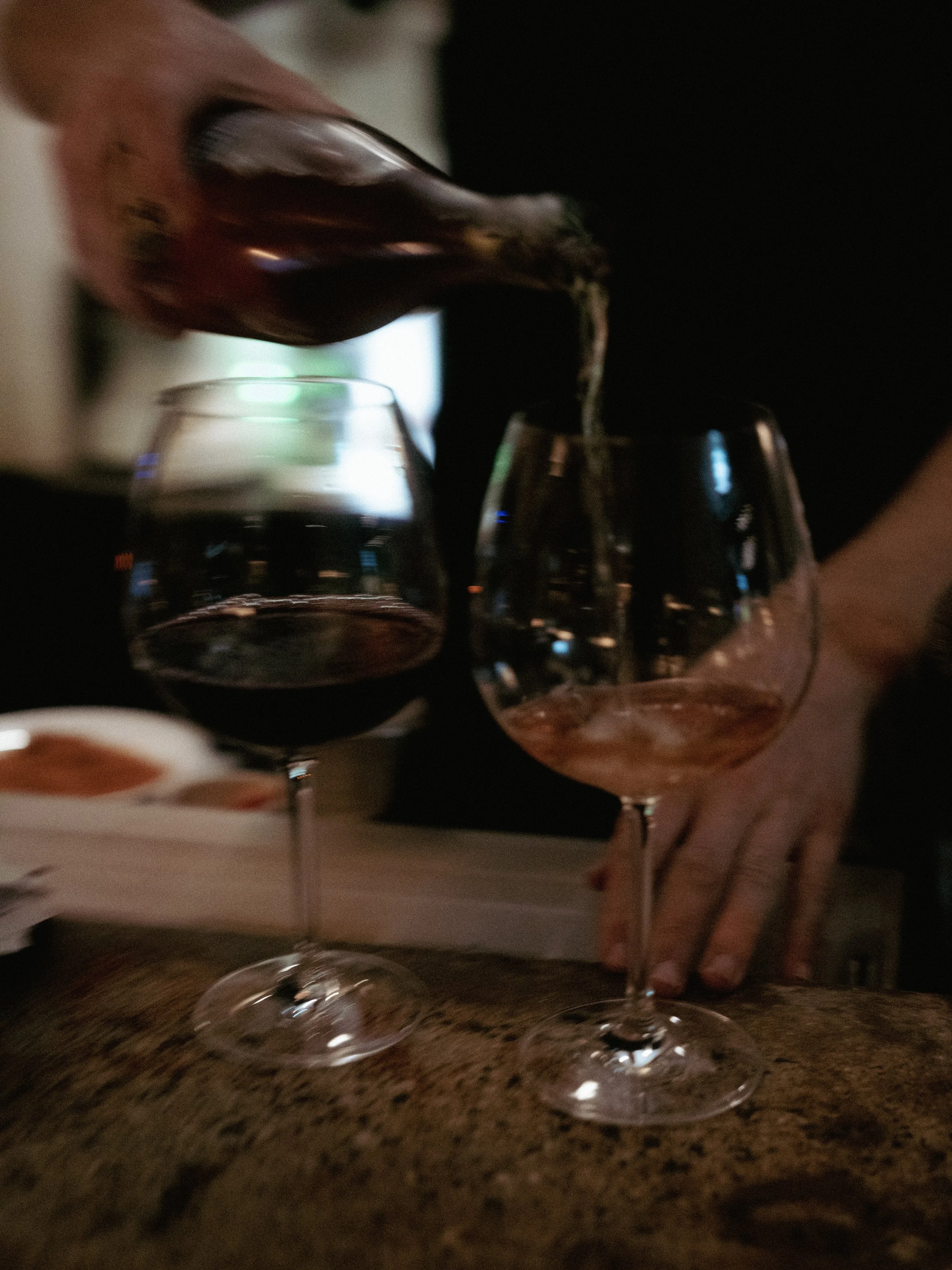 A person is pouring red wine from a bottle into a wine glass on a wooden table, with another similar glass nearby.