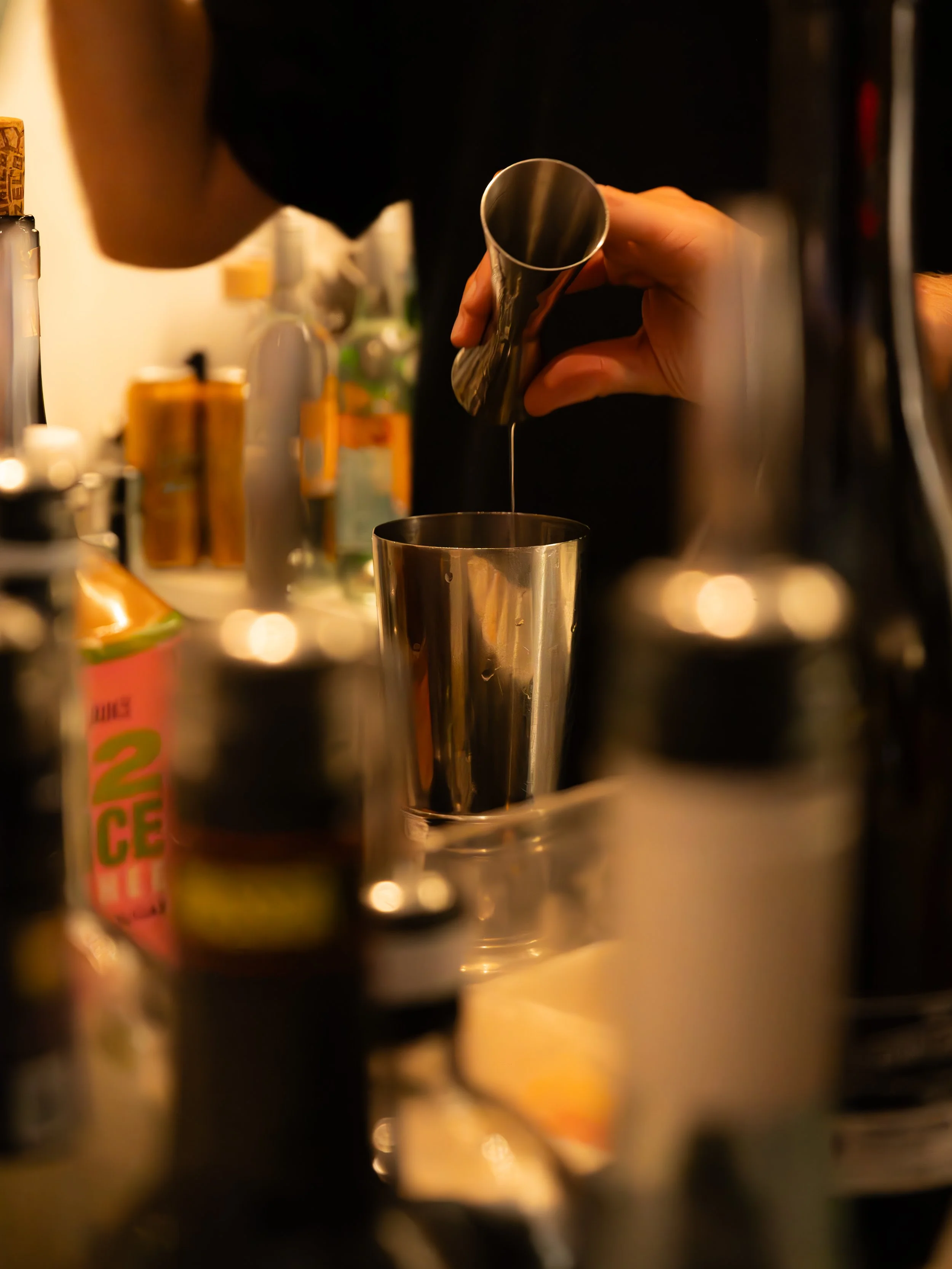 Bartender pouring liquid into shaker with various bottles in foreground