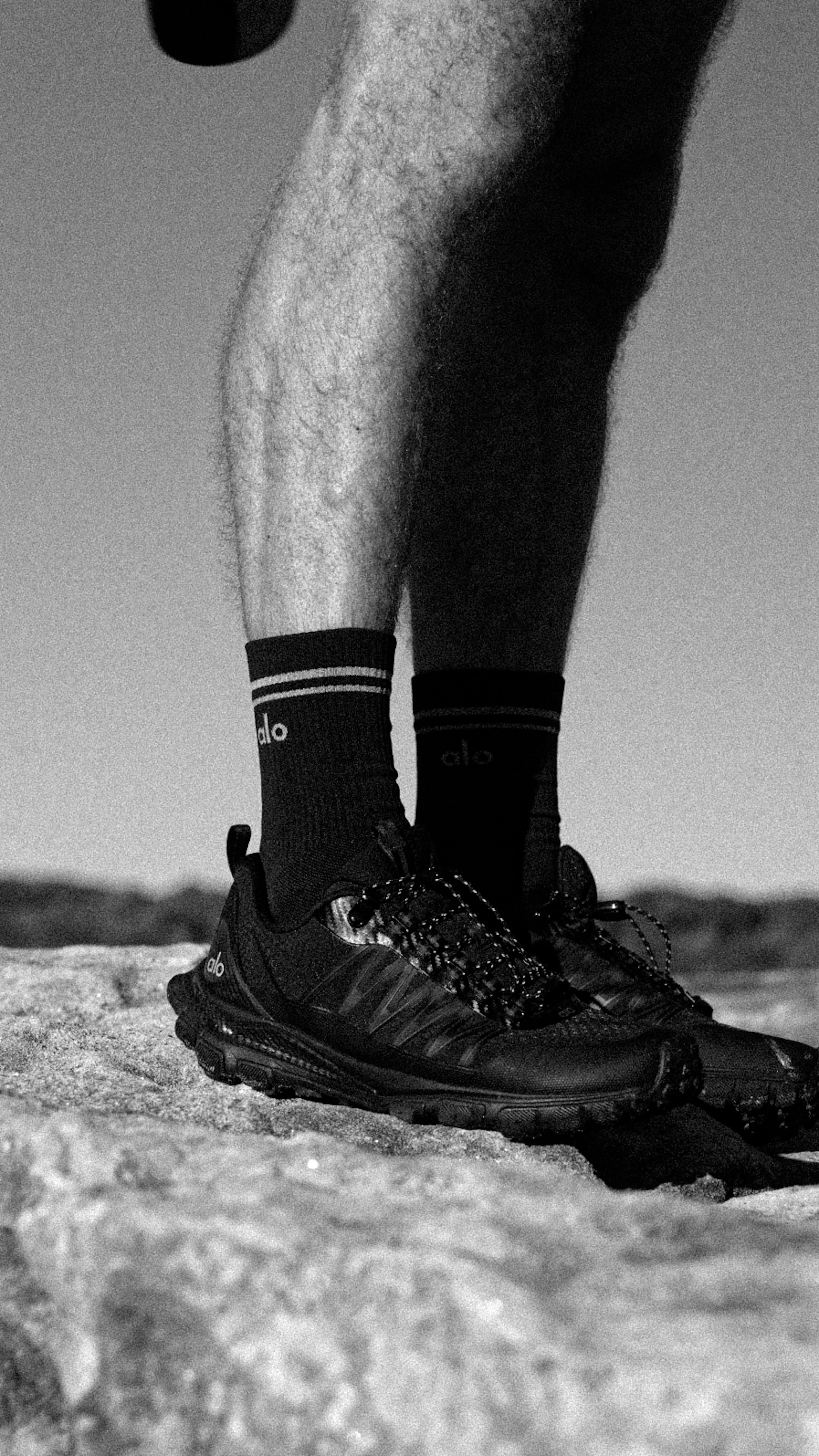 Close-up of a person's lower legs and feet wearing black athletic shoes, black socks with white stripes, standing on sandy ground.