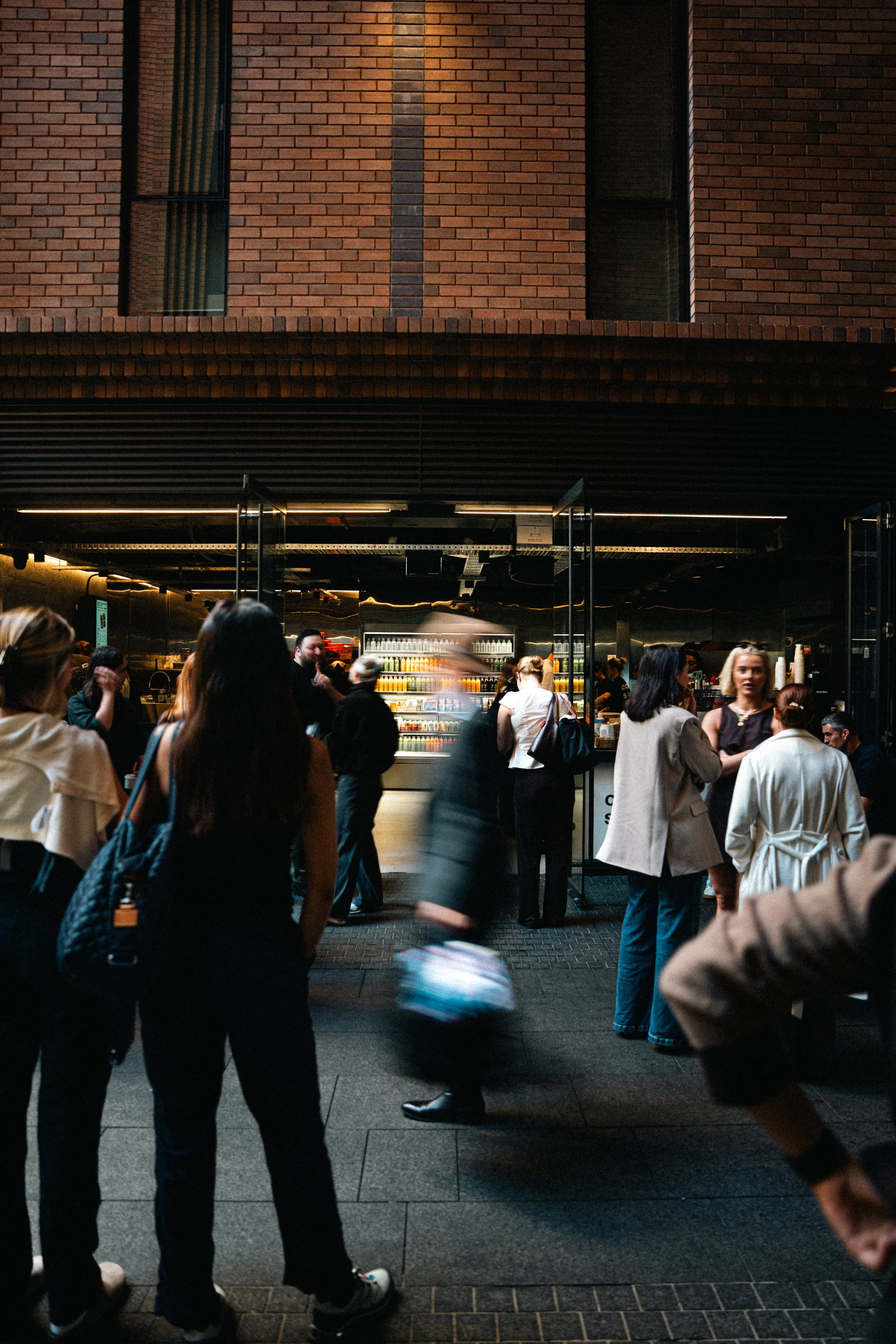 People gathering outside a brick building with an open entrance, some engaged in conversation, and shelves visible inside with drinks.