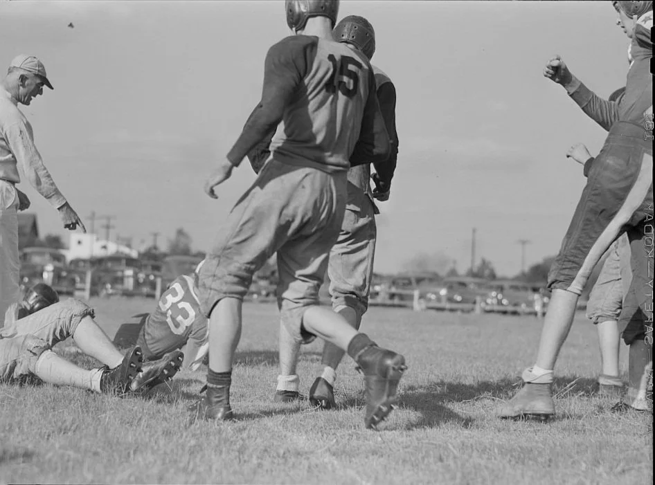 Football players on a field during a game, some standing and some on the ground, with one player on the ground tackled and others watching or celebrating.