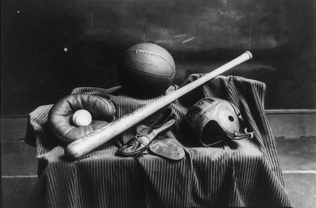 Black and white photograph of vintage baseball equipment on a draped cloth, including a baseball, a baseball glove, a baseball bat, and a catcher's mask, with a black background.