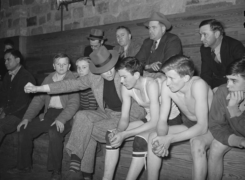 Group of young boys and adult men sitting on a wooden bench, with some boys shirtless and looking down or to the side, in what appears to be a vintage setting.