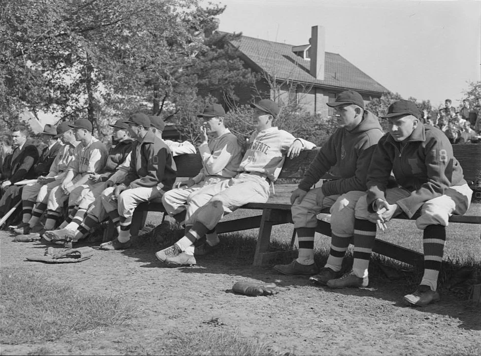 Young boys in baseball uniforms sitting on a bench at a baseball field, some holding bats, with a house and trees in the background.