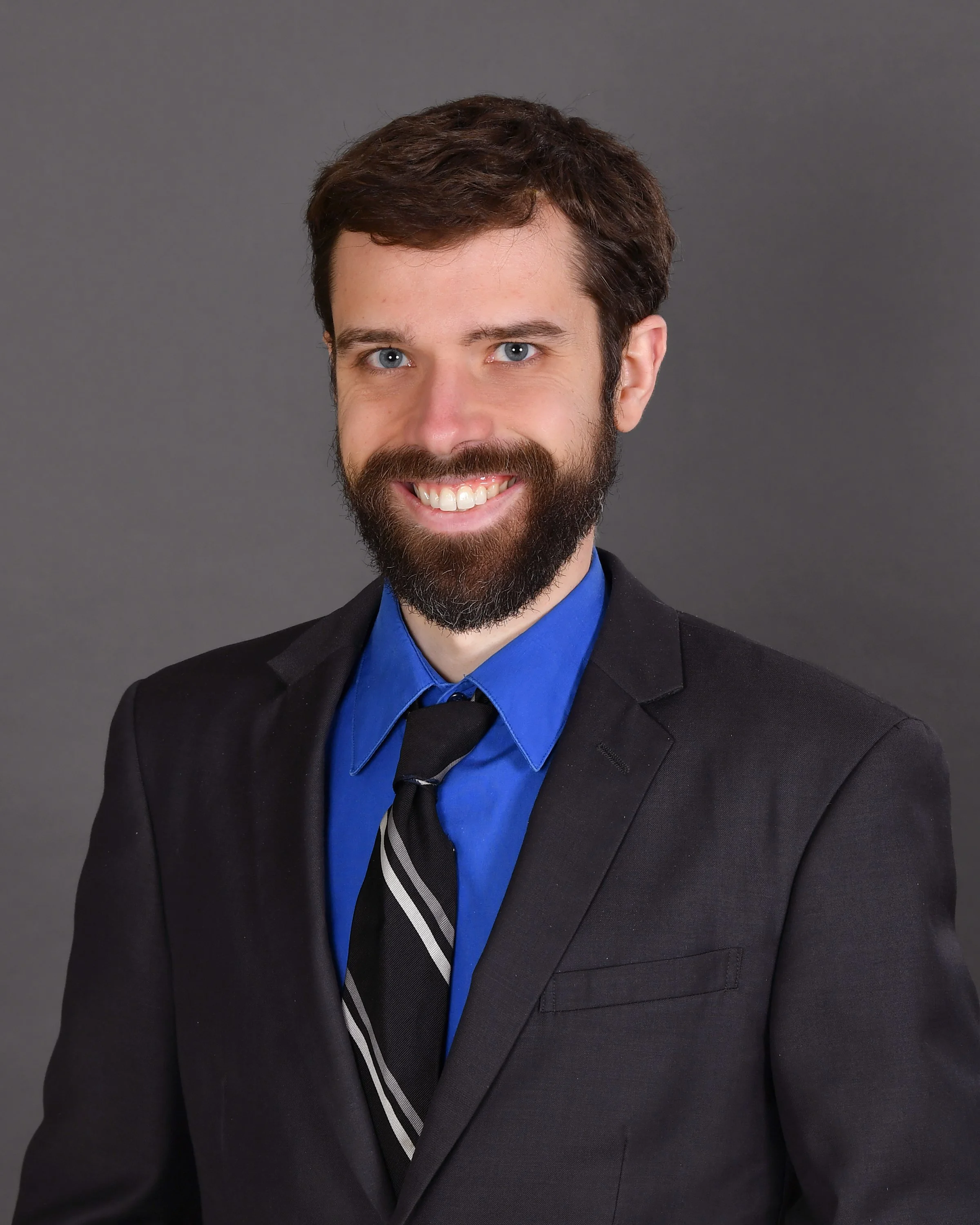 A man with brown hair and a beard, wearing a dark suit, blue shirt, and striped tie, smiling in front of a gray textured background.