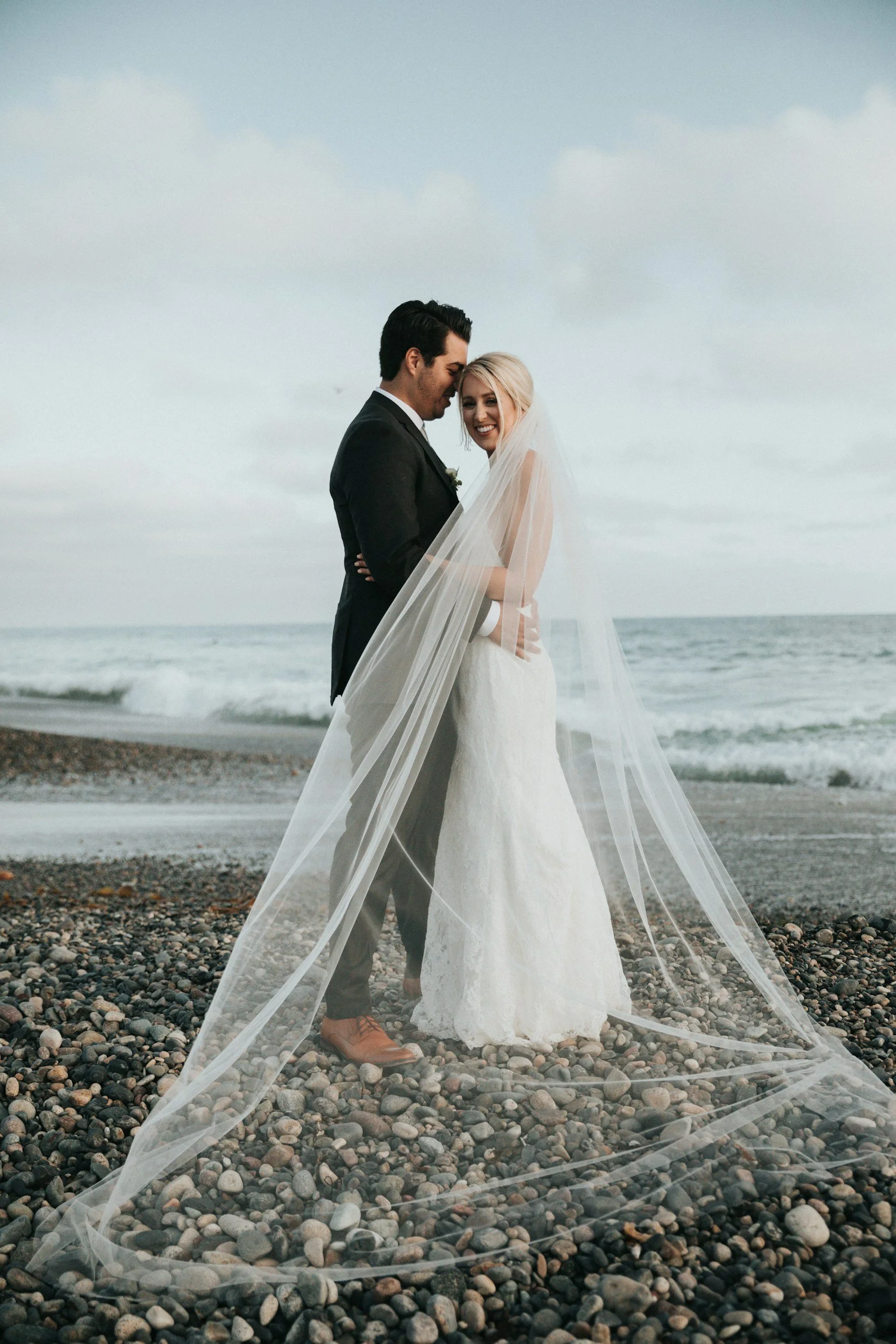 Bride and groom embracing on a pebbled beach, with the ocean in the background and a flowing veil.
