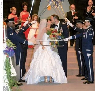 Bride and groom kiss under a military saber arch at a wedding ceremony, surrounded by military personnel in uniforms and guests in formal attire.