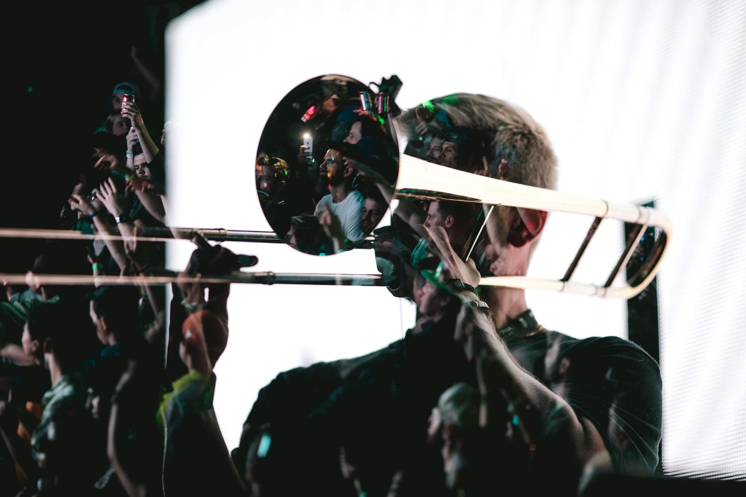A musician plays a trombone on stage, with a reflection of the audience in the instrument's bell, showing crowd members taking photos and watching.