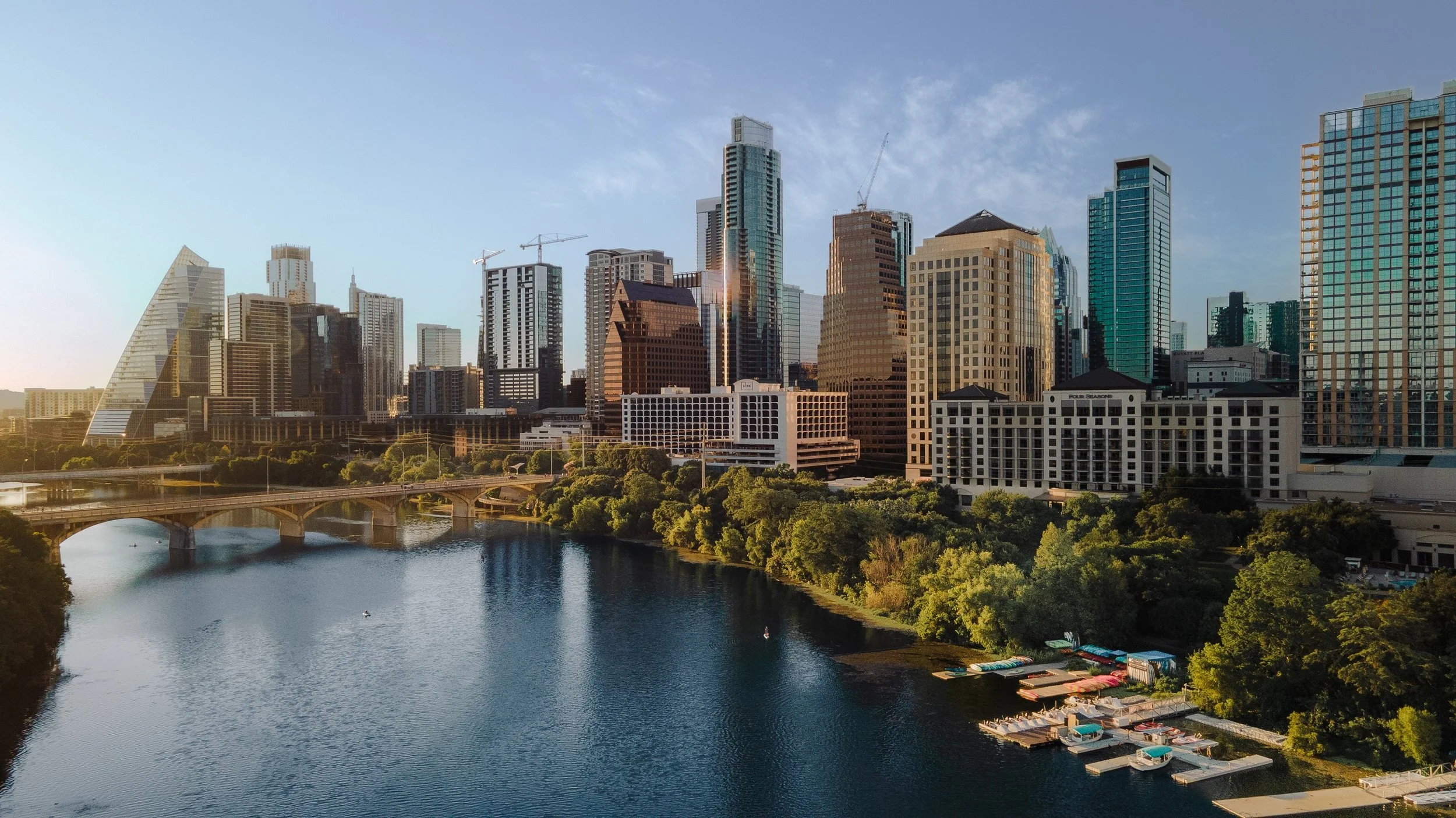 Skyline of a modern city with high-rise buildings and a river in the foreground, boats, and a bridge, under a partly cloudy sky.