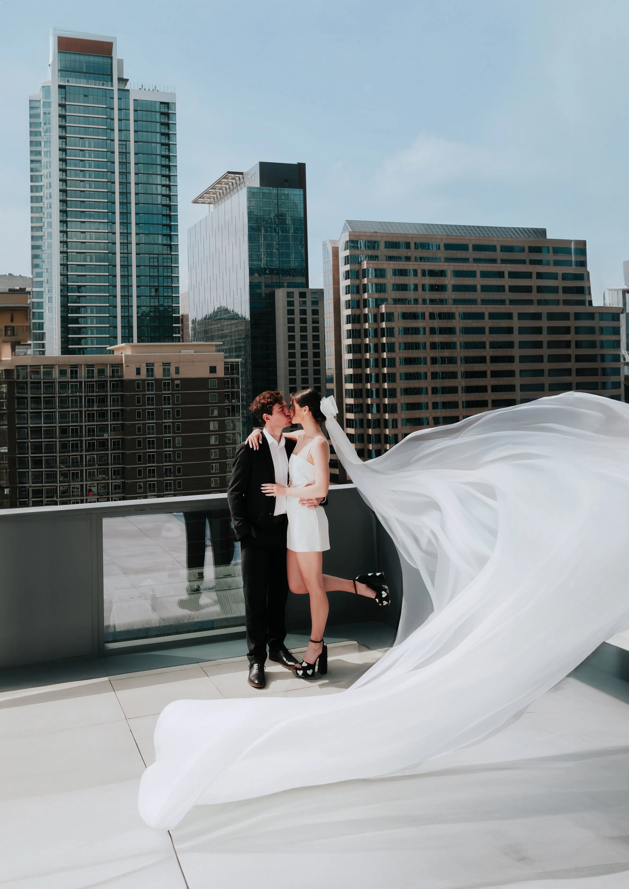 A couple kissing on a rooftop terrace in a city, with a flowing white dress and high heels, surrounded by tall modern buildings.