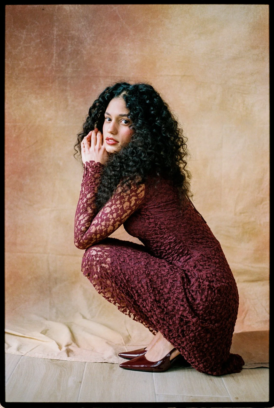 A woman with long curly black hair squatting down, wearing a maroon lace dress and high heels, against a neutral textured background.