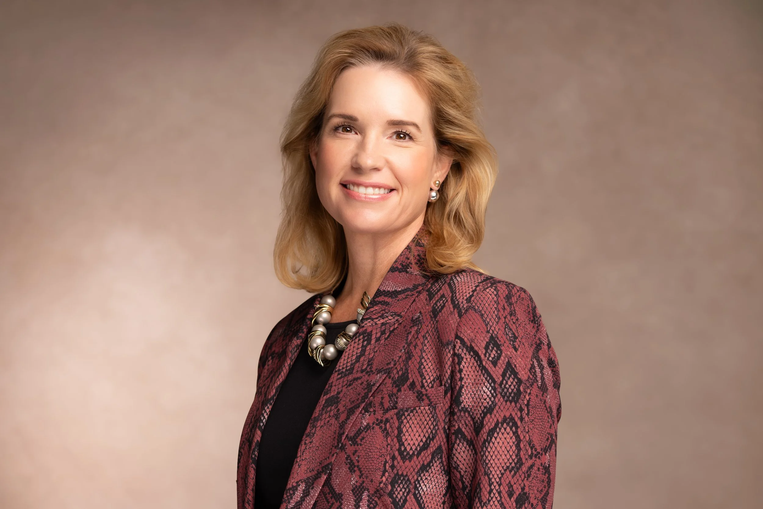 A smiling woman with shoulder-length blonde hair wearing a patterned blazer, black top, and pearl necklace, posing against a plain beige background.