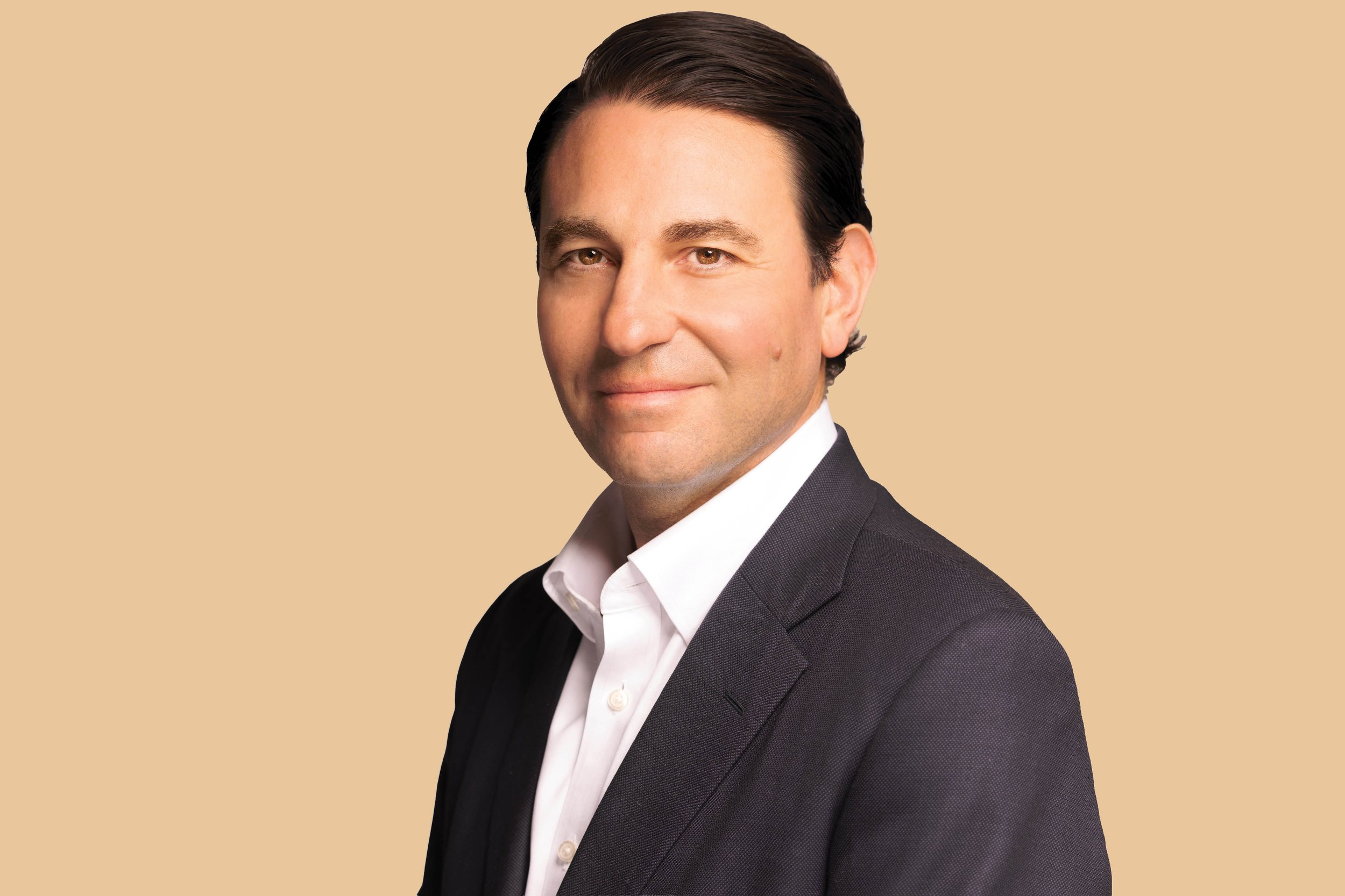 Professional man with dark hair and light skin wearing a black suit and white shirt, standing against a beige background.