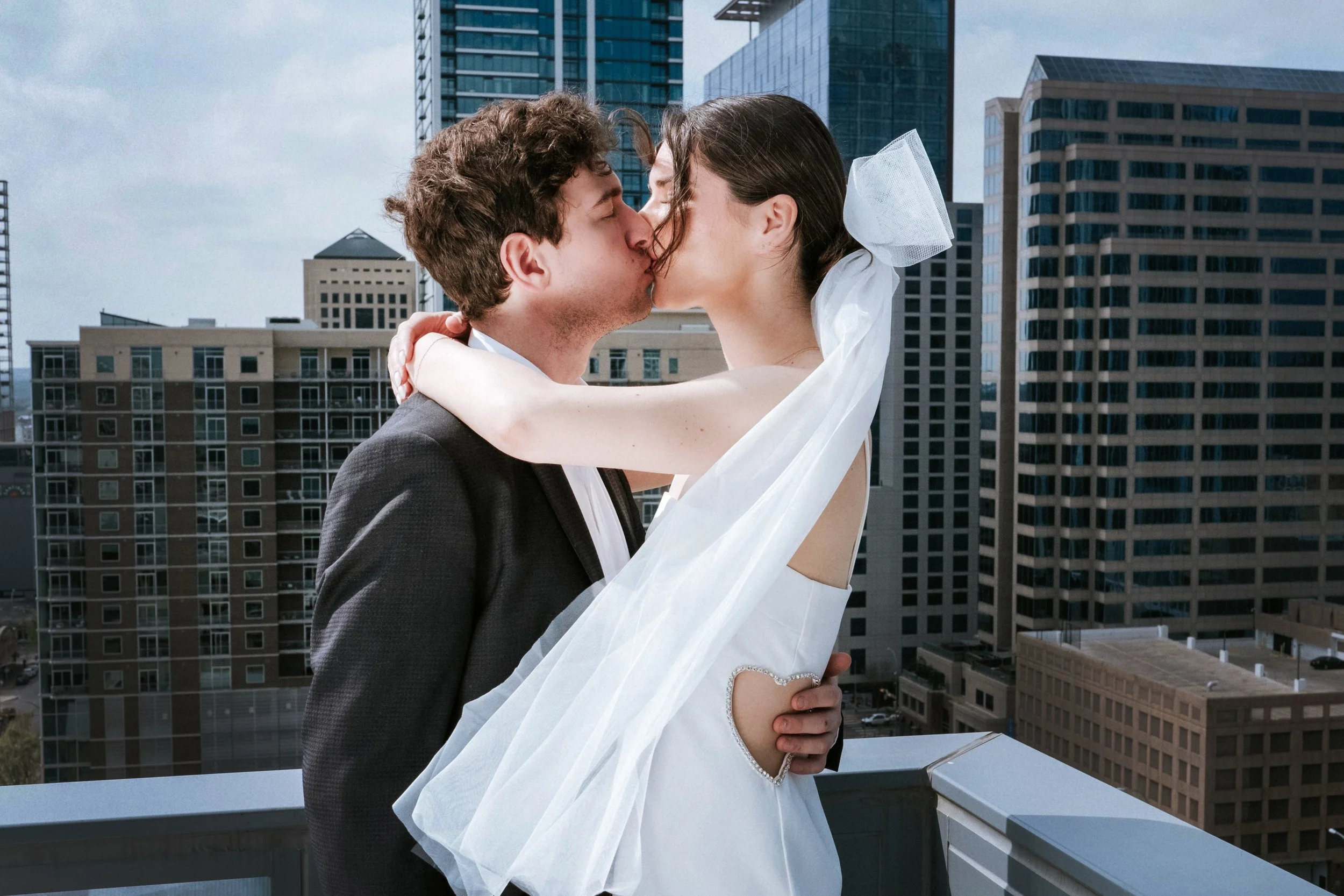 A couple kissing on a rooftop with a city skyline in the background, the woman wearing a white dress and veil, and the man in a dark suit.