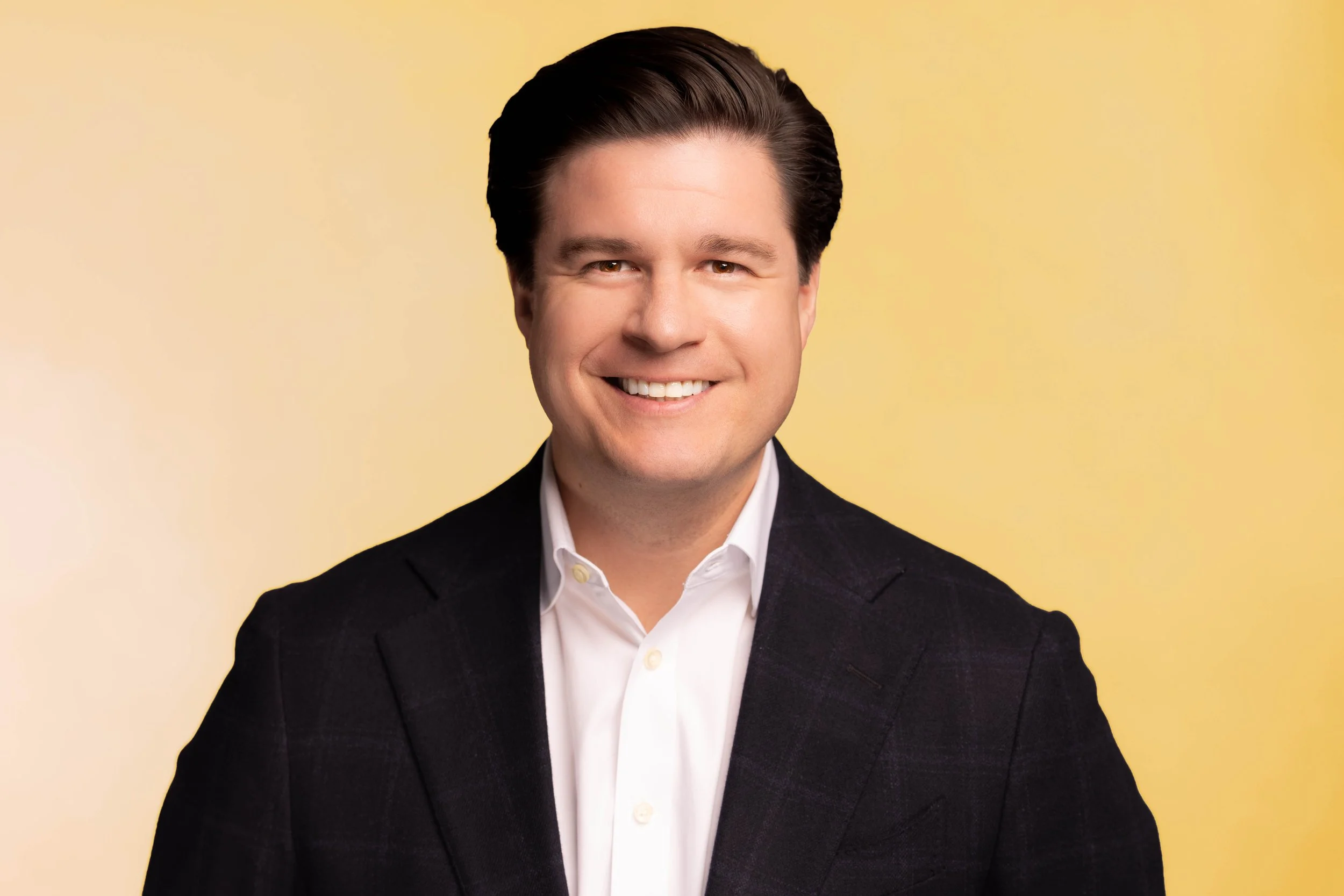 A portrait of a smiling man in a black suit and white shirt against a yellow background.