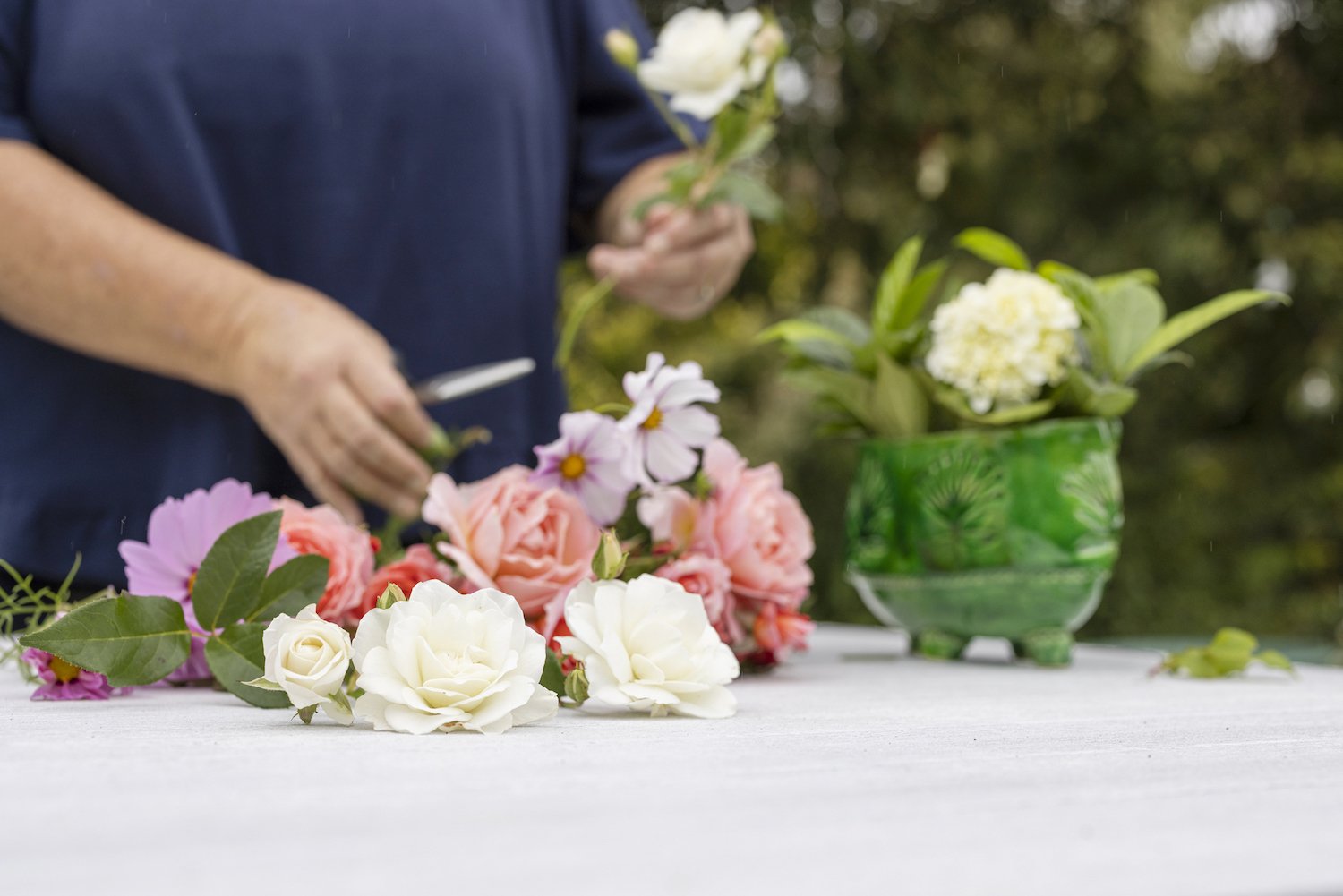 Person arranging flowers on a table outdoors with a green vase of white flowers in the background.