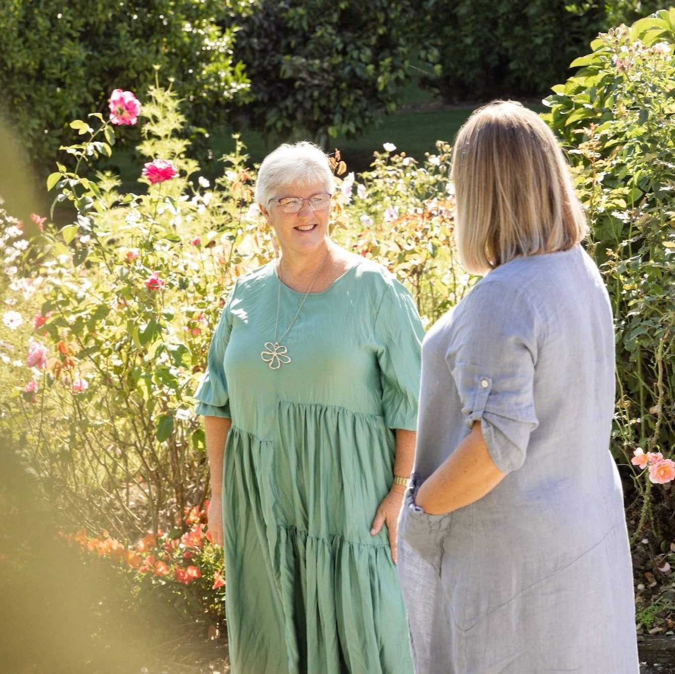 Two women sharing a conversation outdoors in a garden with blooming flowers, one elderly woman with glasses and short white hair wearing a green dress, and another woman with shoulder-length blonde hair wearing a light-colored shirt.