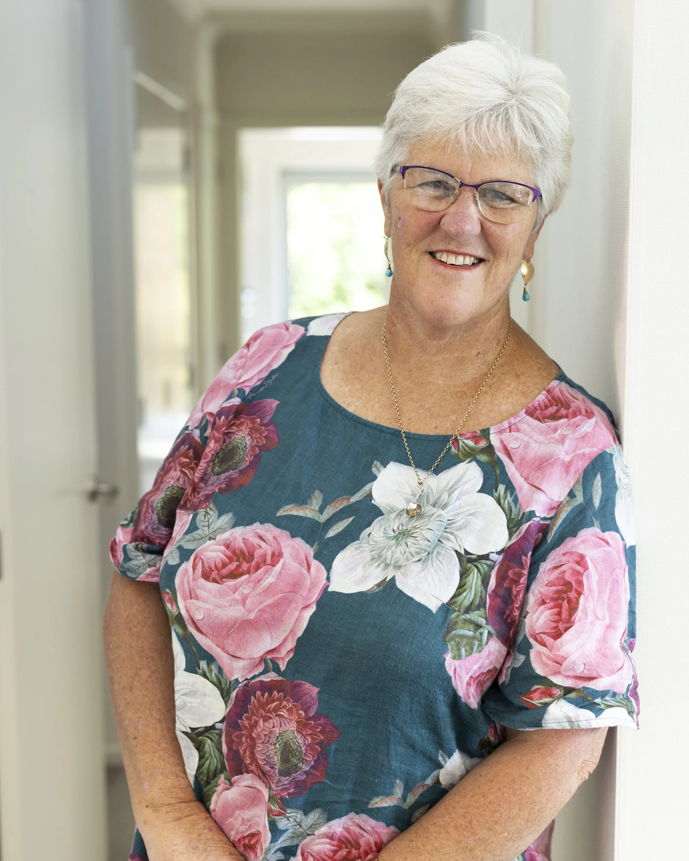 Smiling elderly woman with white hair, glasses, and turquoise jewelry standing indoors near a white wall with natural light.