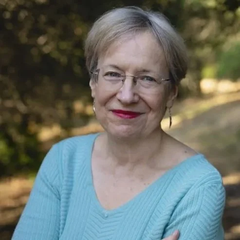 Portrait of an older woman with short blonde hair and glasses, wearing a light blue sweater, standing outdoors in a natural setting.