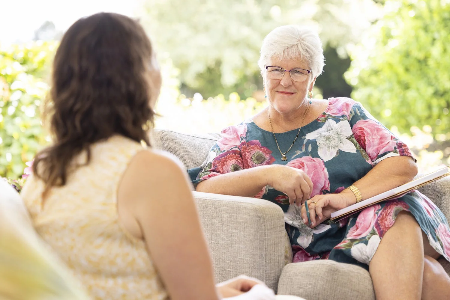 An elderly woman with glasses and a floral dress talking to a younger woman with brown hair in a sleeveless top, seated outside on a couch with a garden background.