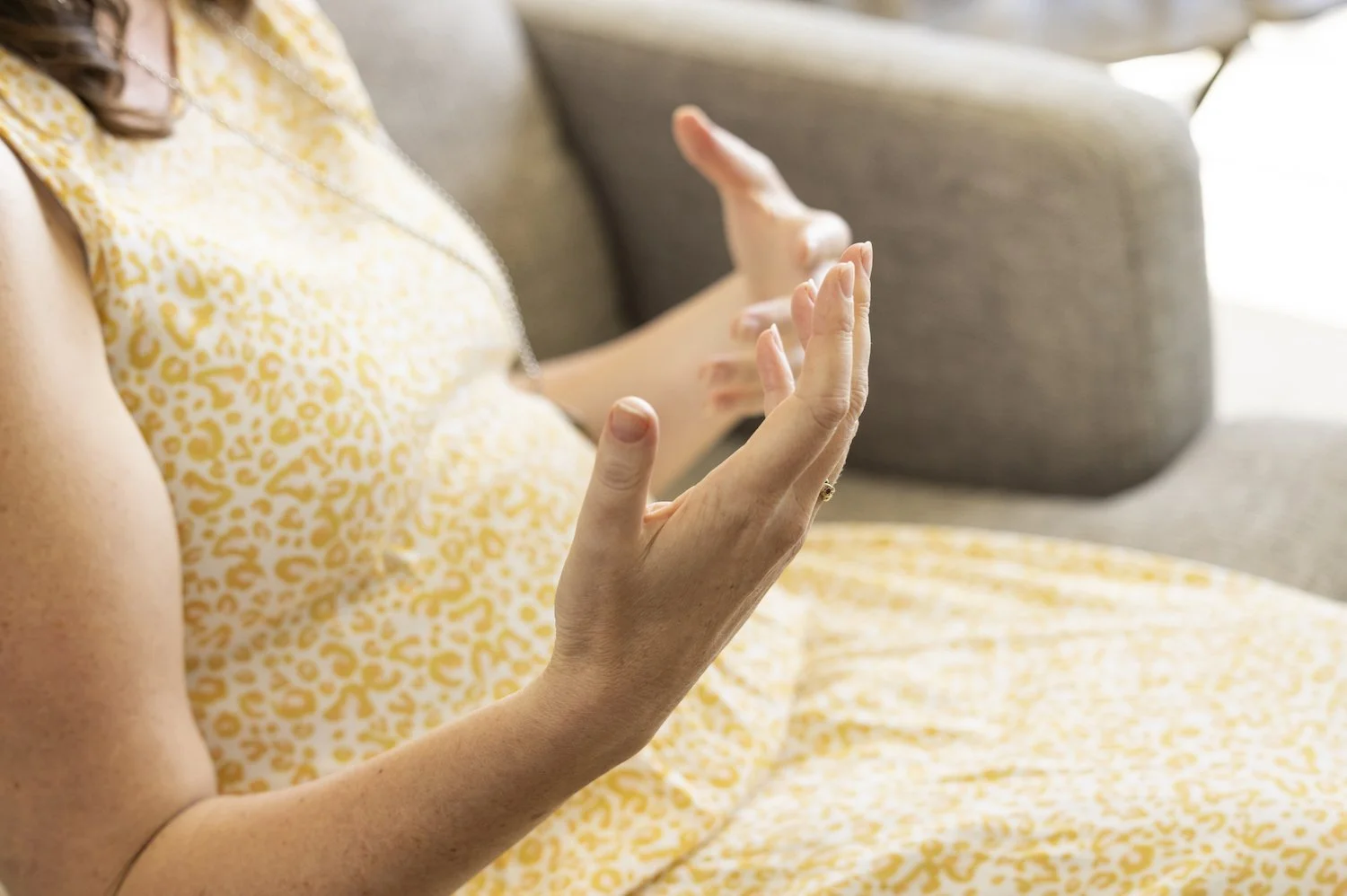 Close-up of a woman with light skin sitting on a couch, wearing a yellow and white animal print dress, with her hands raised and palms facing outward.