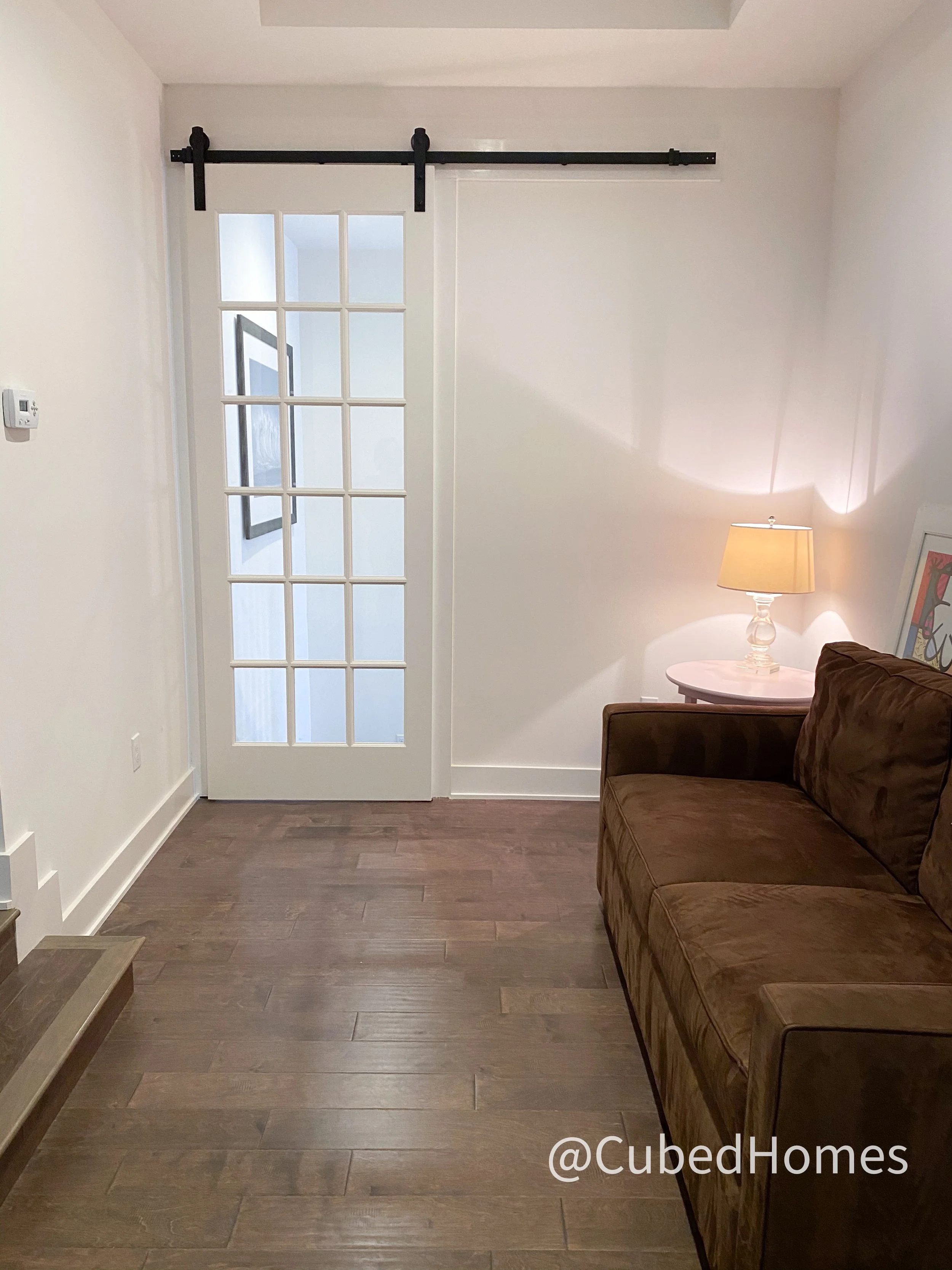 Living room with dark wood floor, brown sofa, white wall with glass-paned door on the left, small oval side table with a lamp on it, artwork leaning against the wall, and a network thermostat on the left wall.