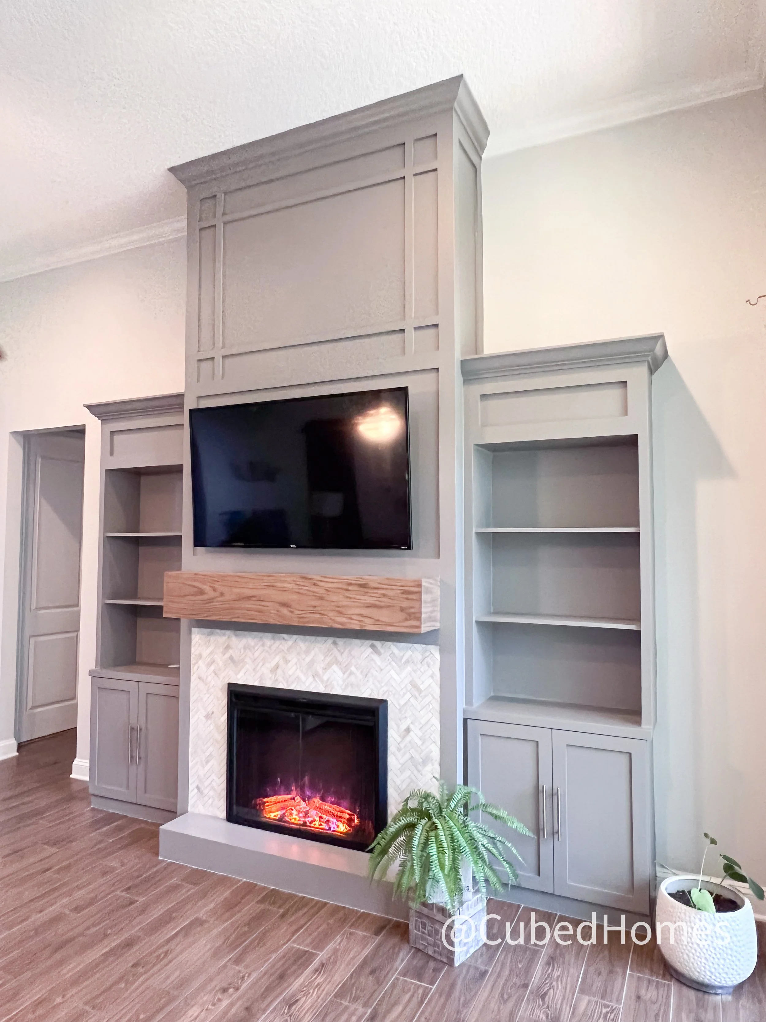 Living room corner with built-in gray bookshelves, a wall-mounted TV above a wooden mantel, a gas fireplace with a tile surround, and potted plants on wooden flooring.