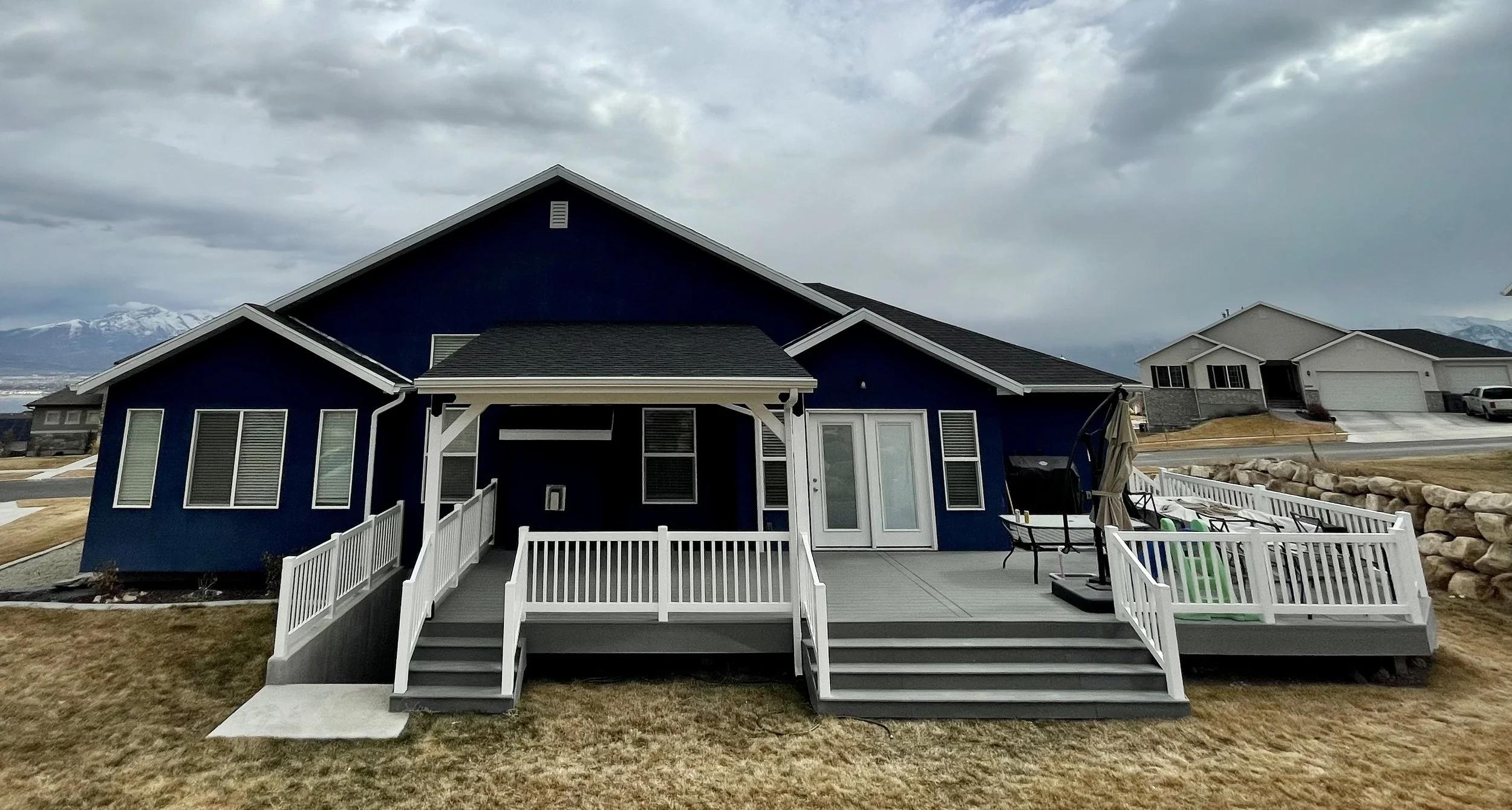 Backyard view of a blue house with white railings and a grey deck, overcast sky, neighboring houses, mountain range in the distance.