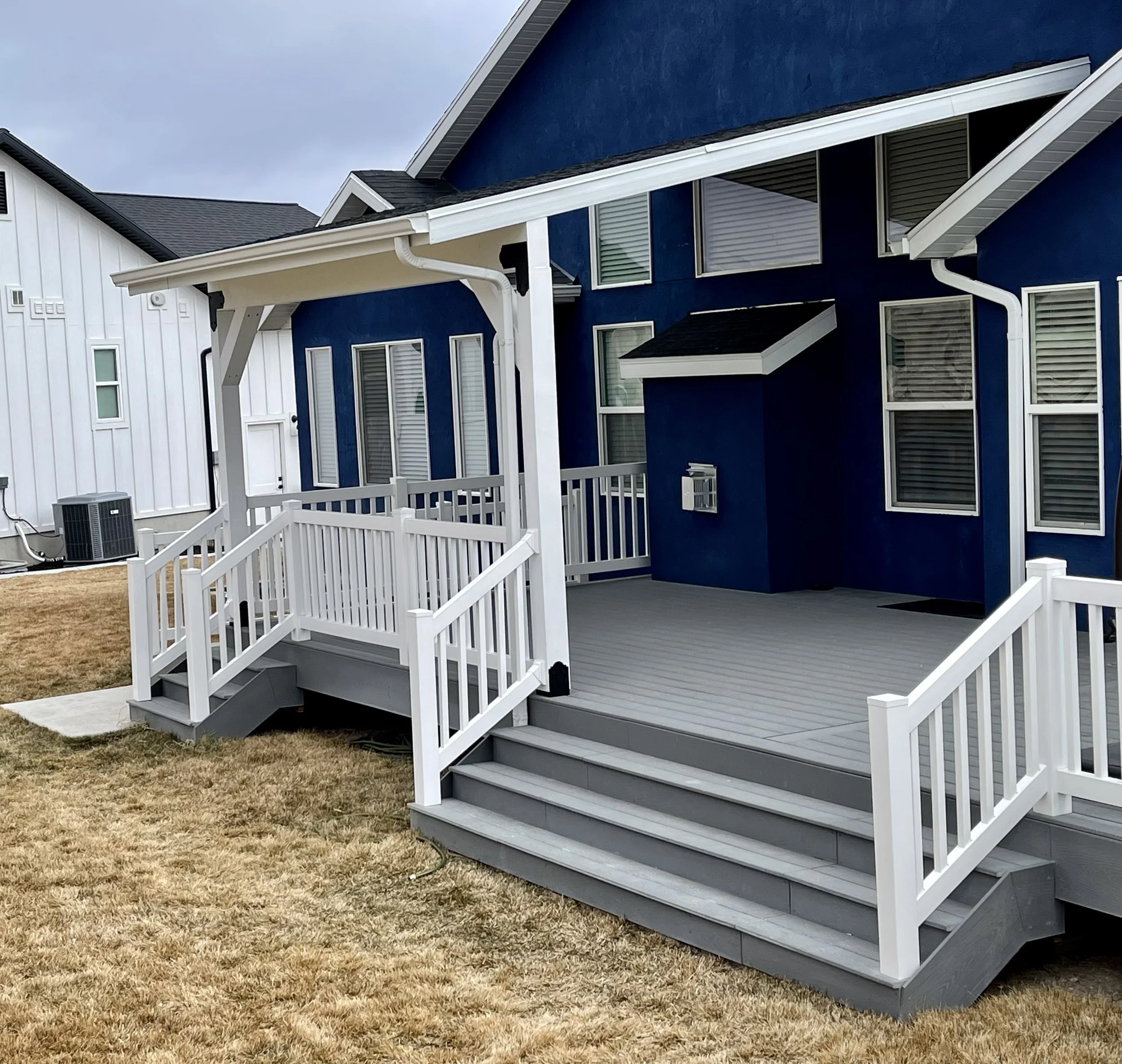 A modern house with a covered porch, white railings, and steps leading down to a lawn with brown grass, featuring a bright blue exterior with multiple windows.