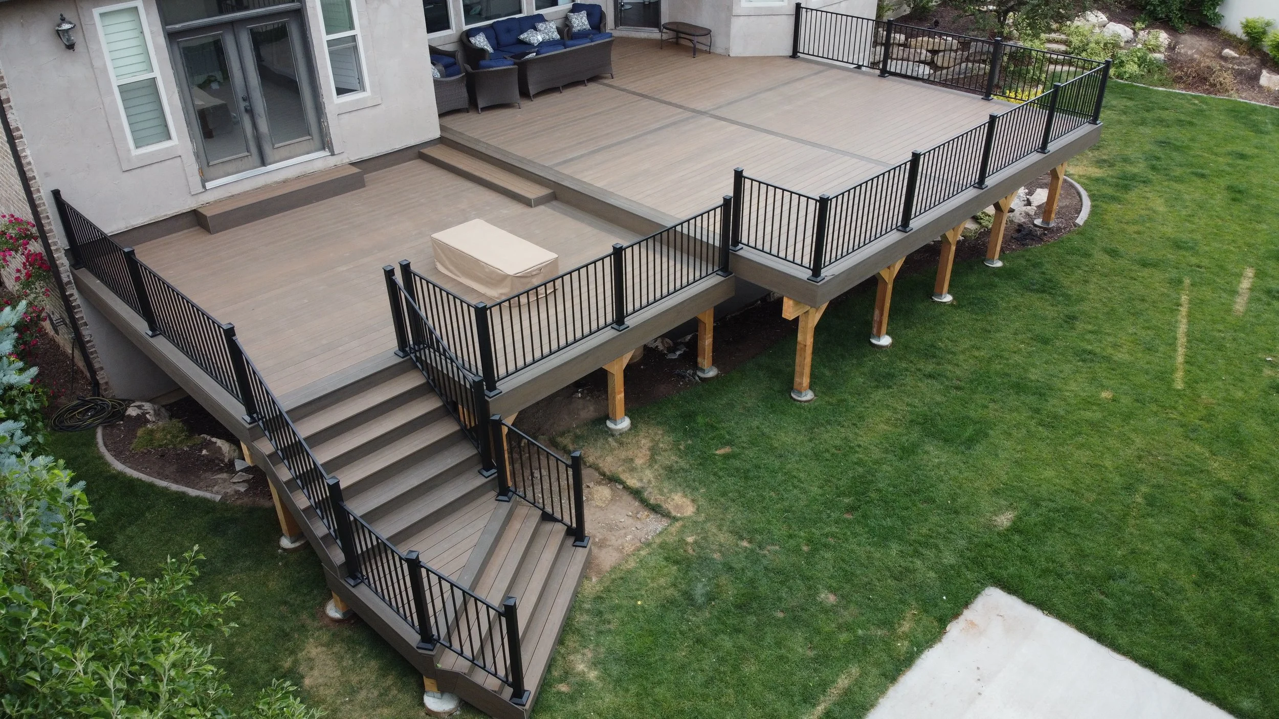 A raised wooden deck with black metal railings and staircases, attached to a house with a sliding glass door, overlooking a green lawn.