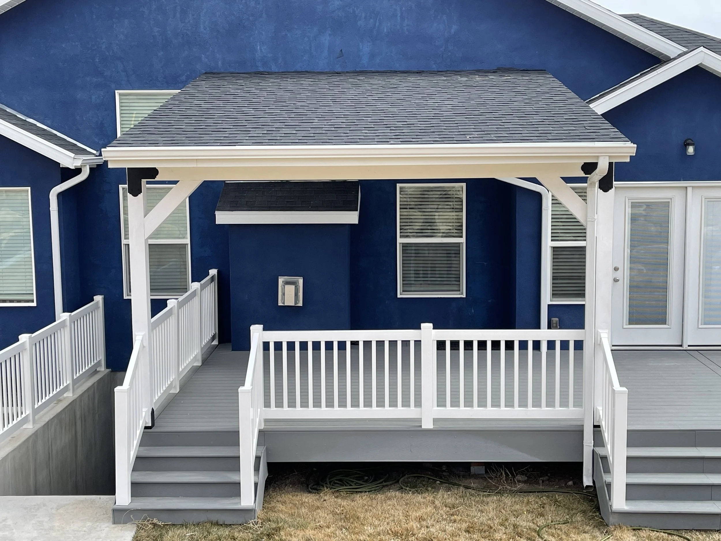 Backyard deck attached to a blue house, with stairs leading down, white fencing, and a covered area with a sloped roof.